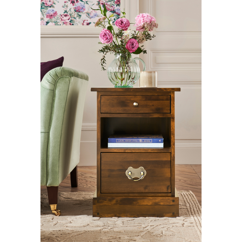 Wooden nightstand with floral arrangement in a room with a chair and decorative wall art.