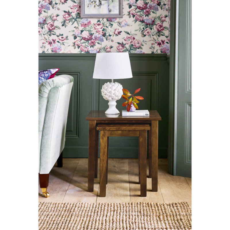 Wooden set of 2 nesting side tables with lamp and decor in a room with floral wallpaper and green paneling.