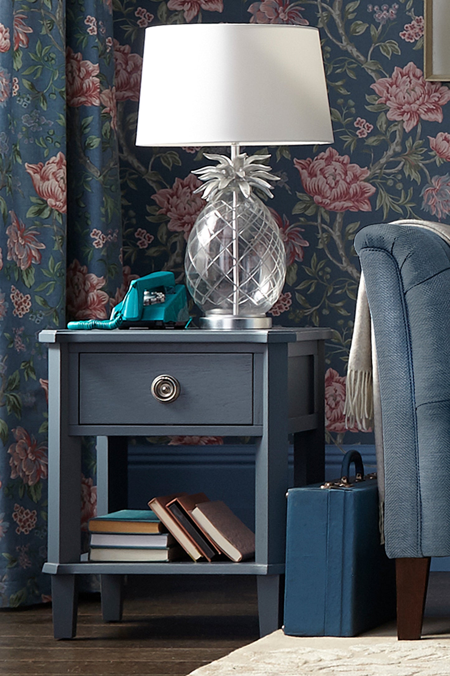 Blue nightstand with lamp, books, and suitcase against floral wallpaper