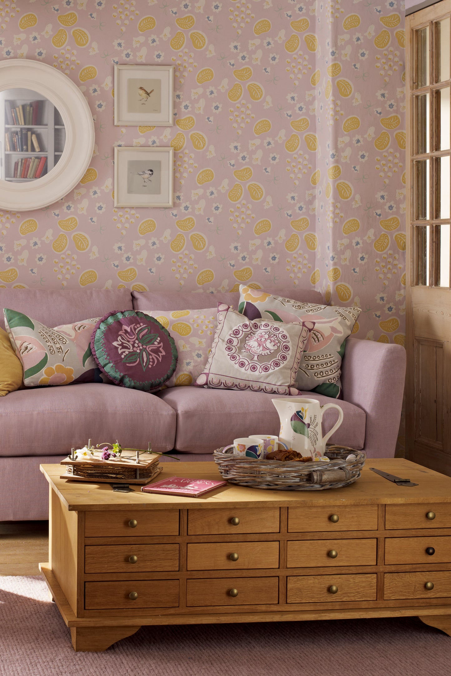 Living room featuring a wooden coffee table with 12 drawers. The room features floral wallpaper, a purple sofa with purple cushions and a wall mirror with 2 pictures hanging up.