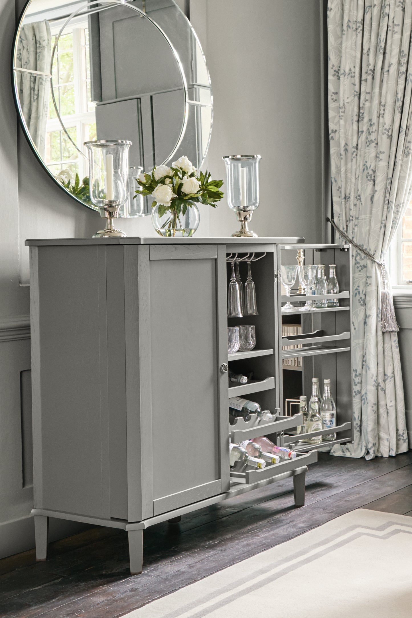 Grey drinks cabinet with open shelves displaying bottles and glasses, floral arrangement, and decorative items in a room with curtains and a mirror.