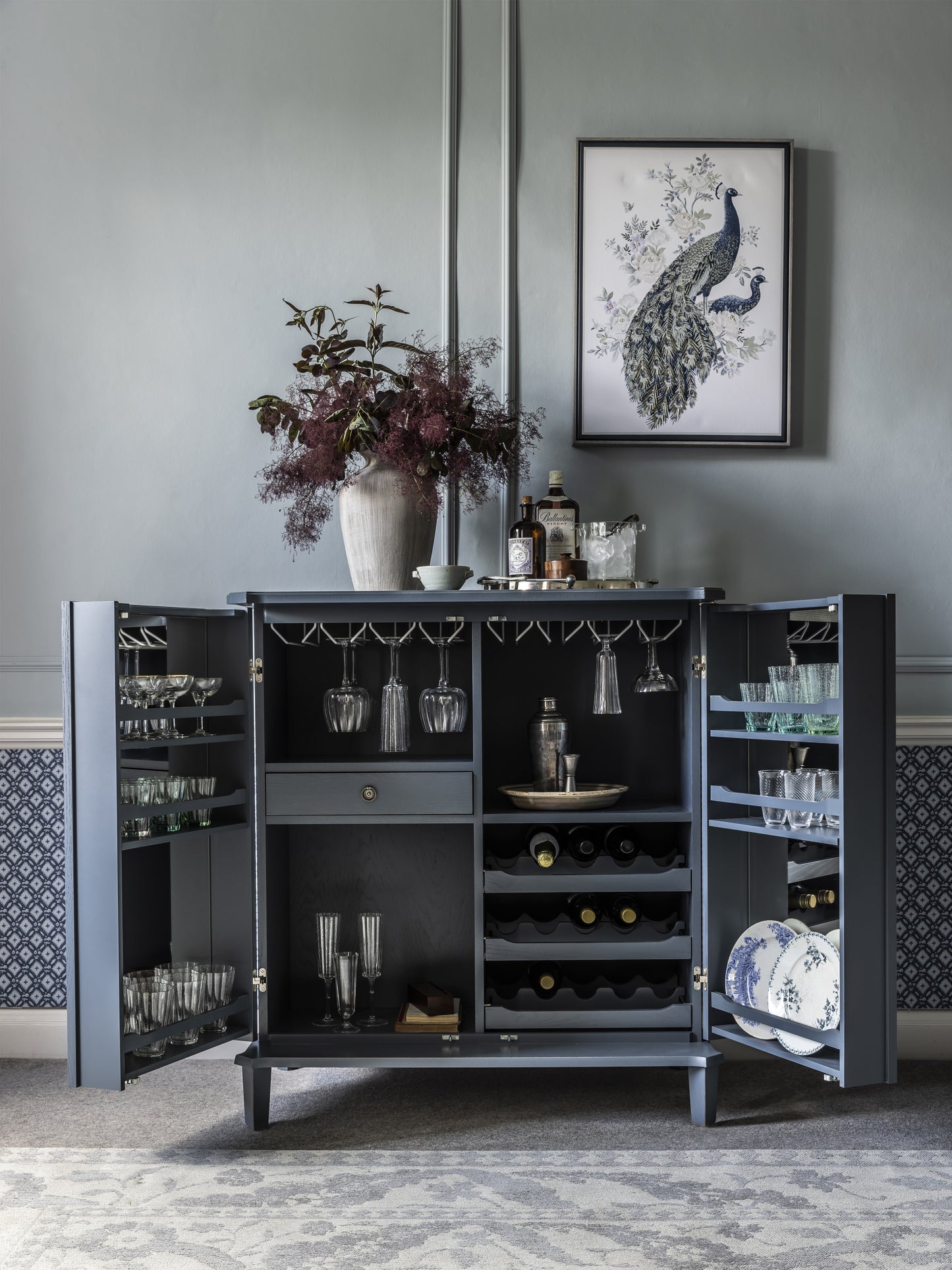 Dark blue drinks cabinet with open doors displaying glasses, bottles, and decor in a room with peacock art on the wall.