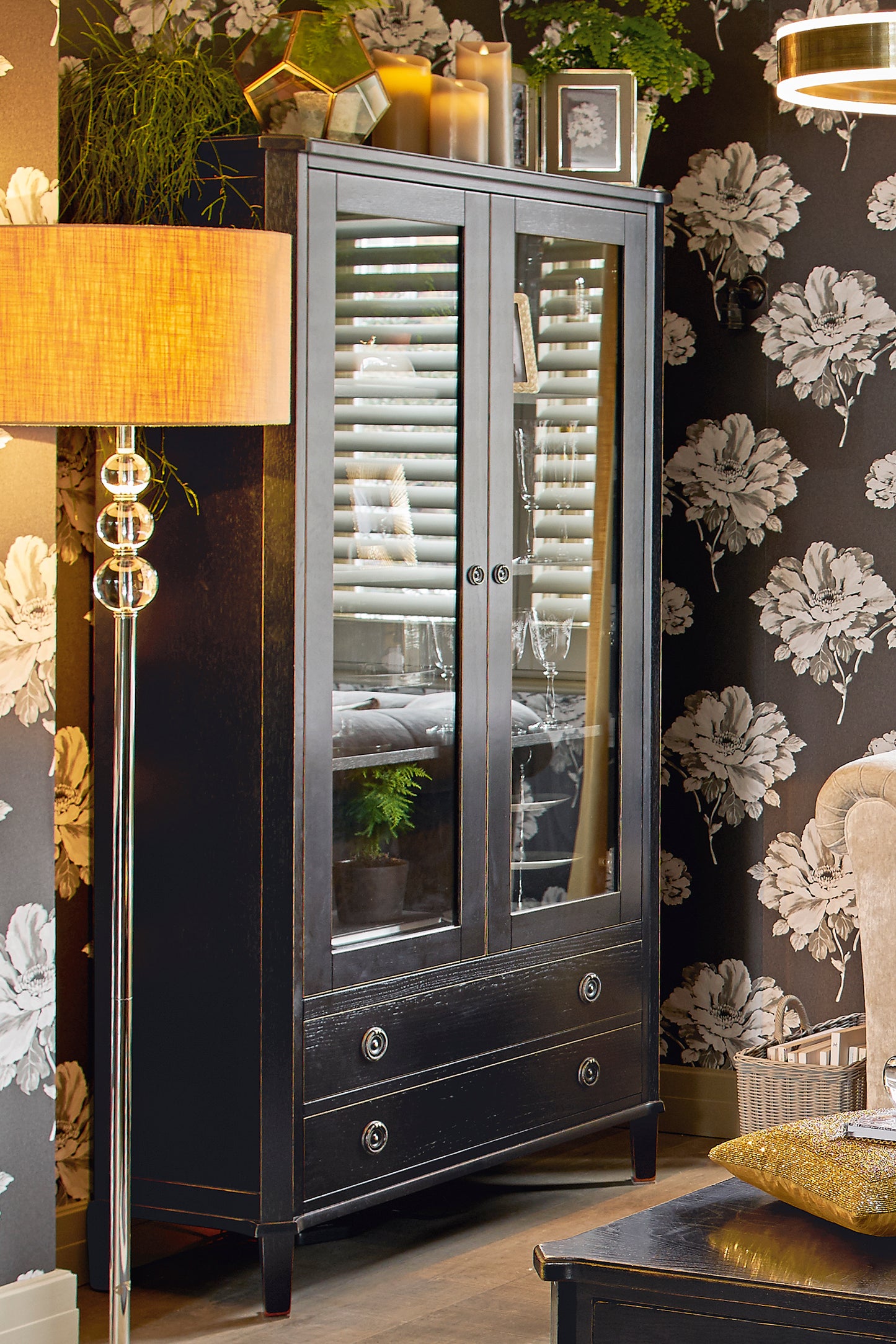Dark wooden cabinet with glass doors in a room with floral wallpaper and a lamp.