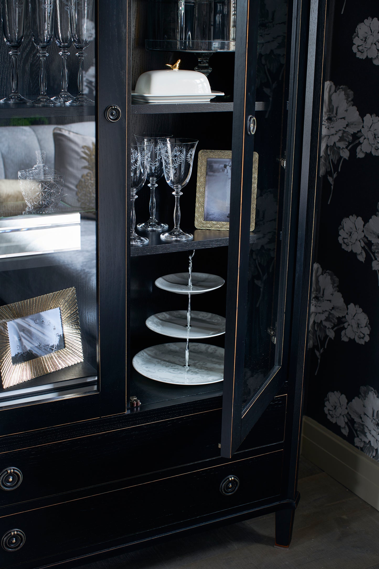 Black display cabinet with glass doors containing decorative items against a floral wallpapered wall.