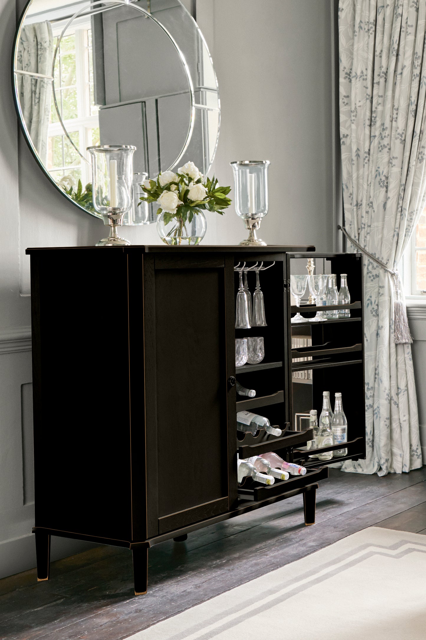 Black drinks cabinet with glassware and flowers in a room with curtains and a mirror.