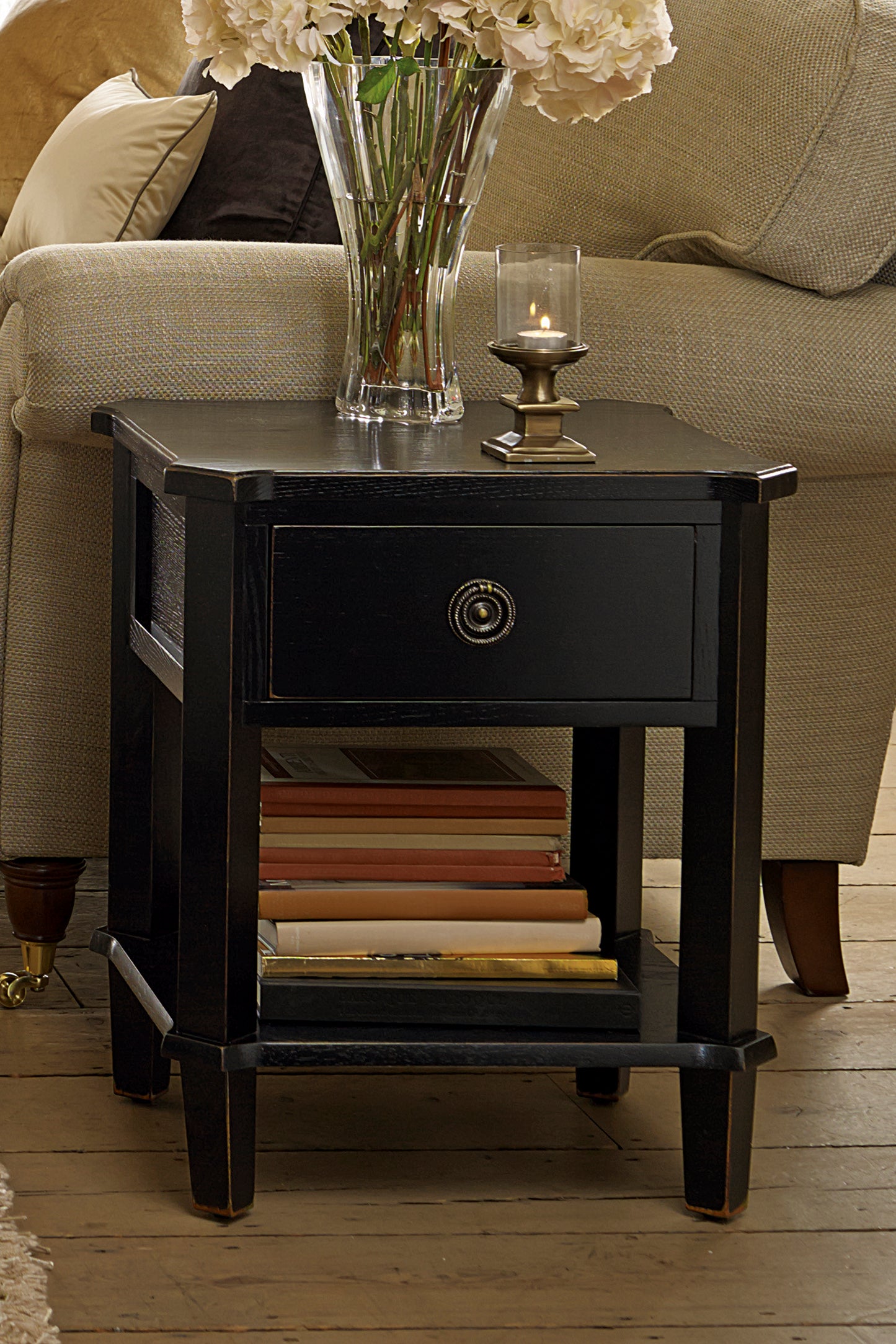 Black end table with a drawer, candle, and vase on a wooden floor next to a beige sofa.
