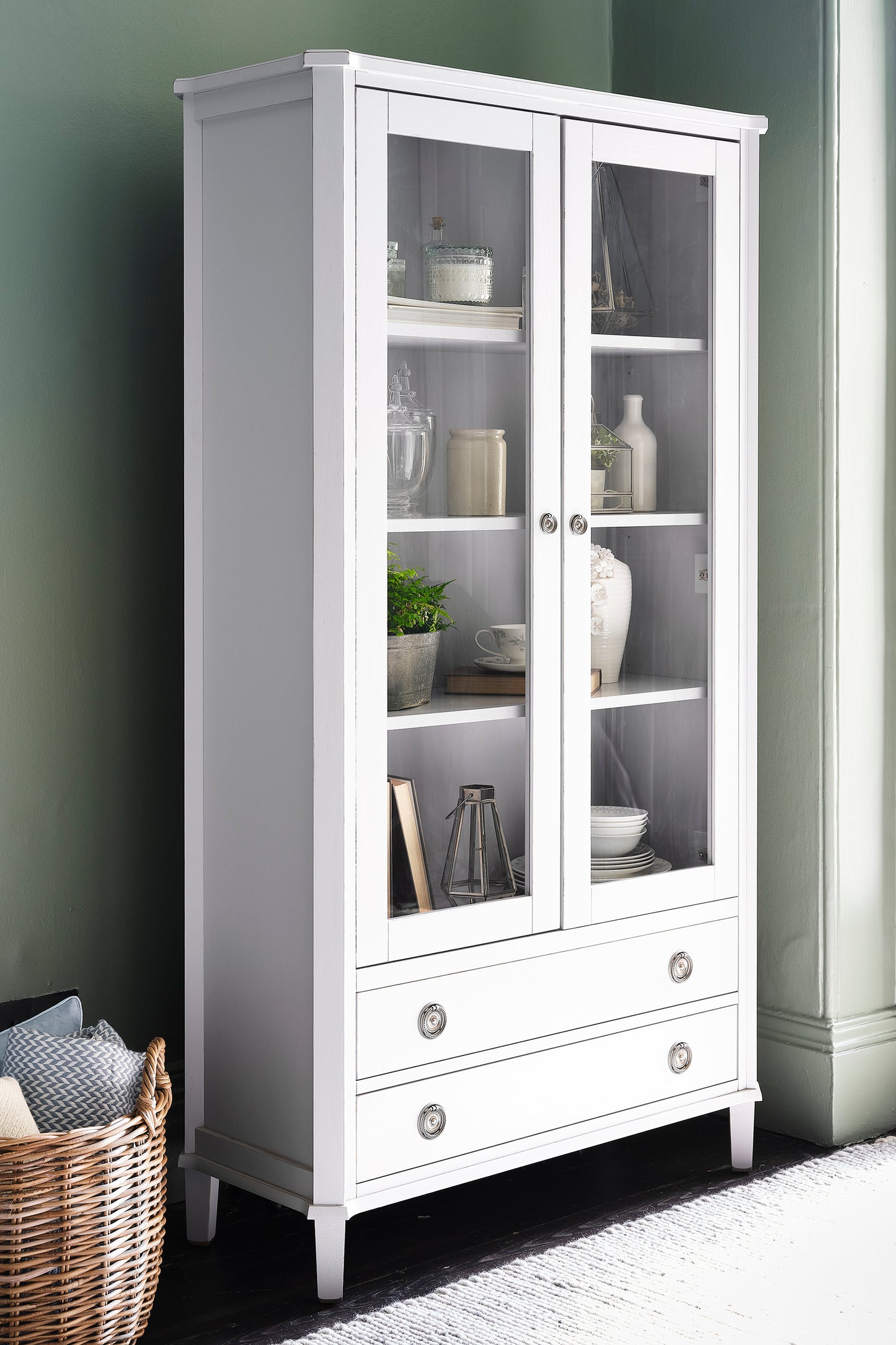 White display cabinet with glass doors and shelves against a green wall.