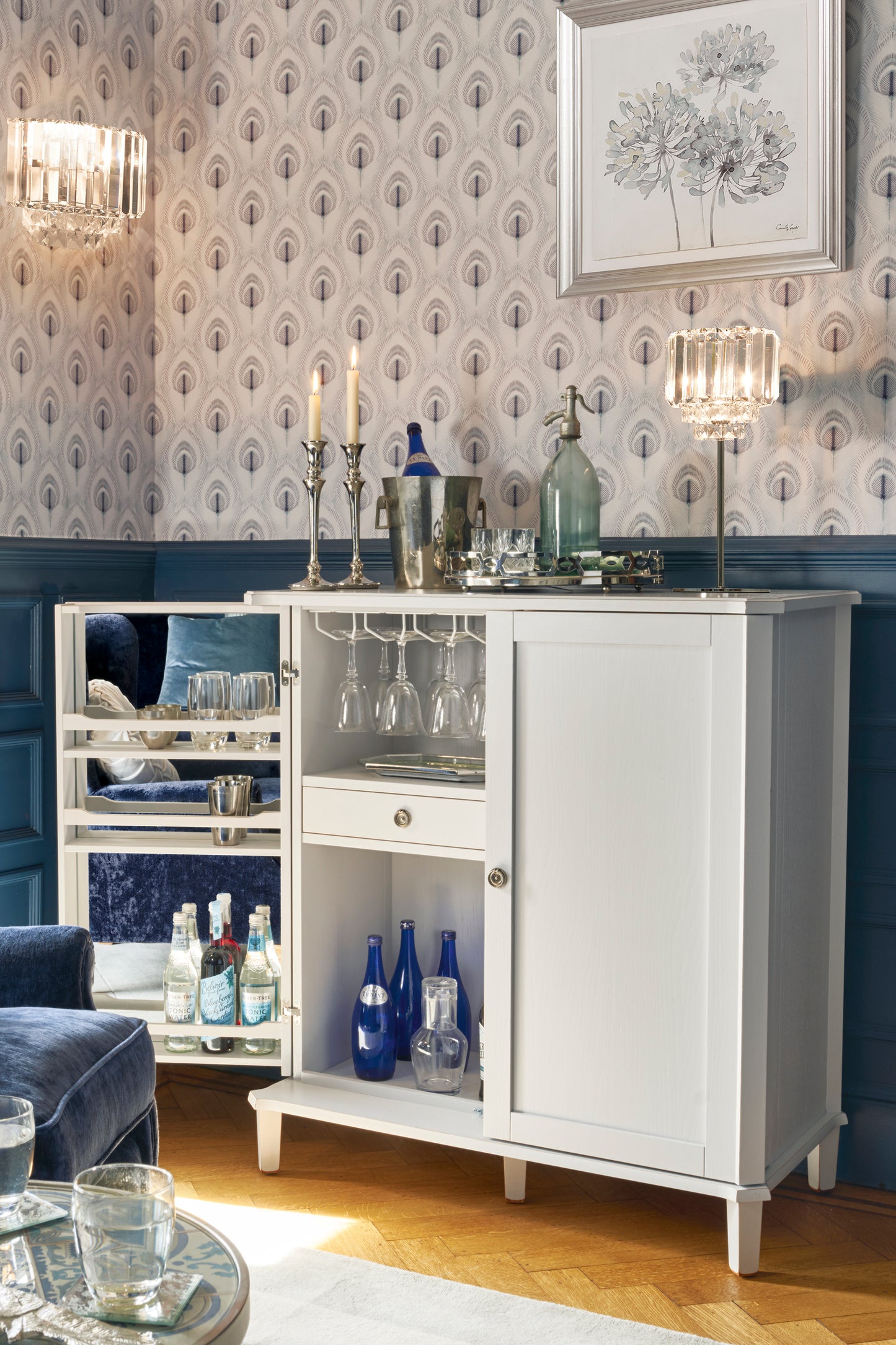 Pale grey drinks cabinet with drinks and glasses in a room with patterned wallpaper.