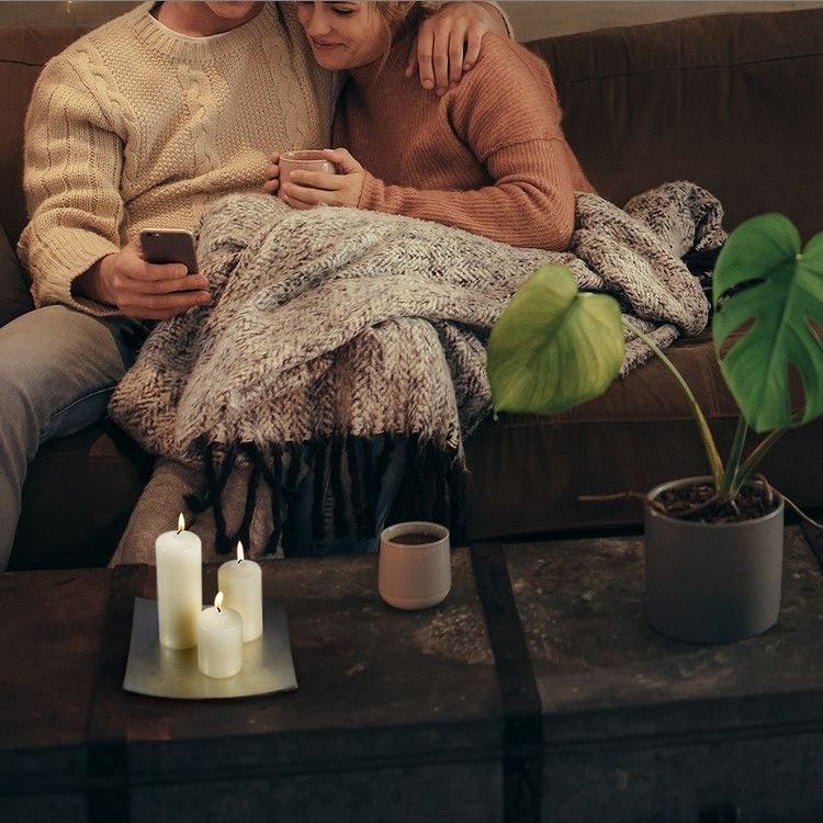 Two people sitting on a couch under a blanket with a candlelit table in front of them.