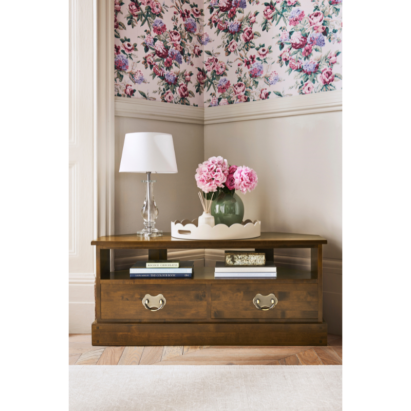 Wooden side table with lamp and flowers against floral wallpaper.