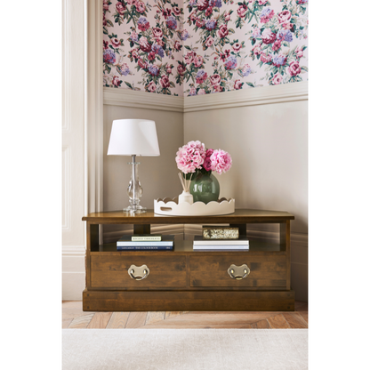Wooden side table with lamp and flowers against floral wallpaper.