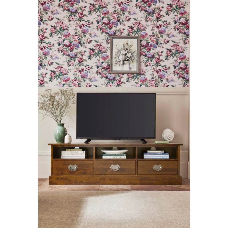 Wooden 3 drawer TV stand with a television and decor in a room with floral wallpaper.
