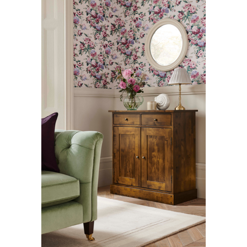 A narrow sideboard with two doors and two drawers, crafted from wood with a brown finish, standing in front of floral wallpaper, wooden cabinet, and green armchair.
