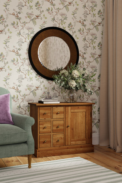A honey-coloured wooden sideboard with one door and six drawers, with a simple traditional design with floral wallpaper and mirror in a room.