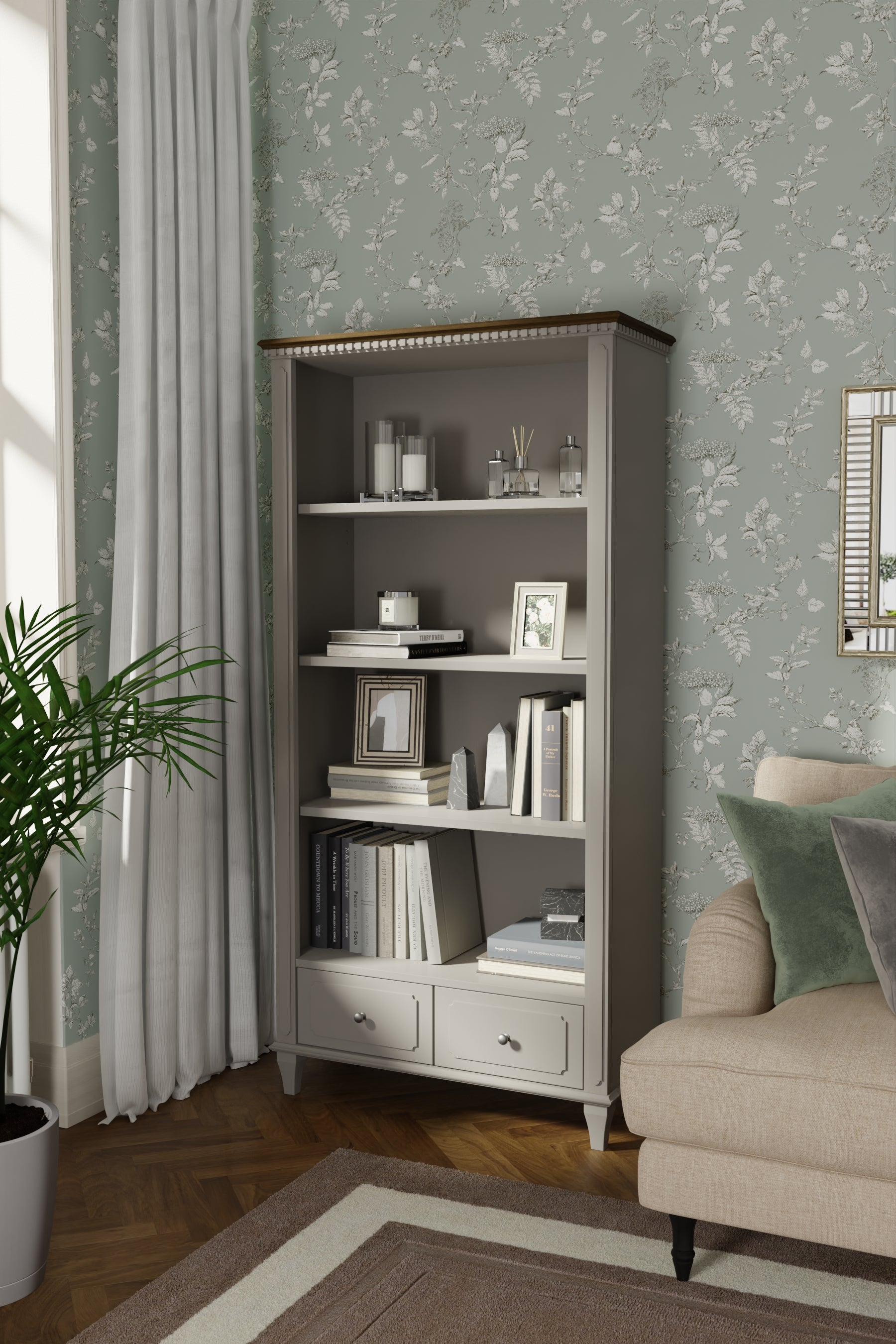 A pale French grey single bookcase with two drawers and wooden details in a room with floral wallpaper, a plant, and a sofa.