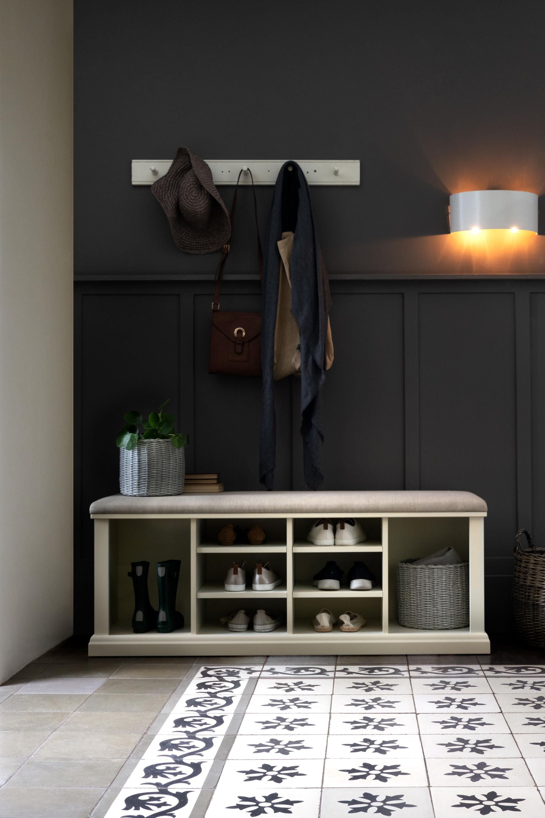A white shoe storage bench with multiple compartments and a cushioned seat on a dark background with coat hooks above and various pairs of shoes in the compartments
