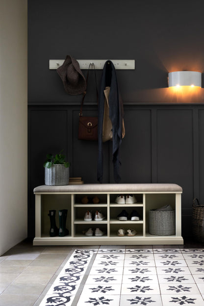 A white shoe storage bench with multiple compartments and a cushioned seat on a dark background with coat hooks above and various pairs of shoes in the compartments