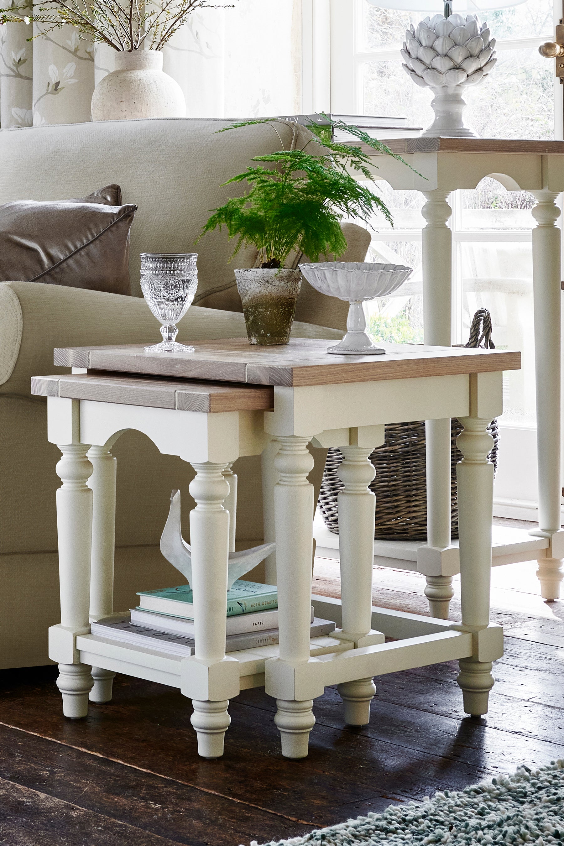 A white and brown nest of two tables with a wooden top and carved legs. Showcased next to a sofa with various ornaments including a plant and a glass