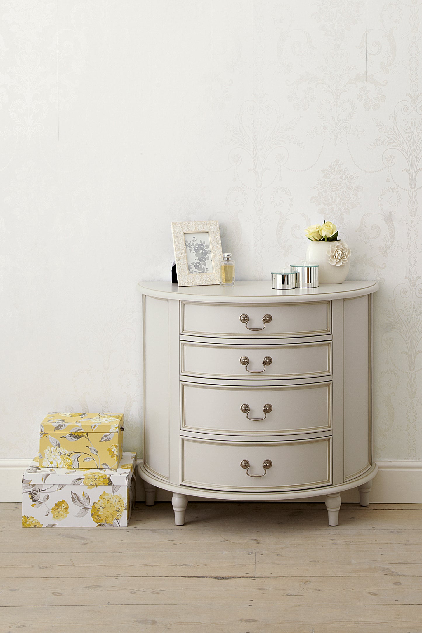 A dove grey 4-drawer half moon chest of drawers with a curved front and traditional metal handles with various ornaments sitting on top and next to it