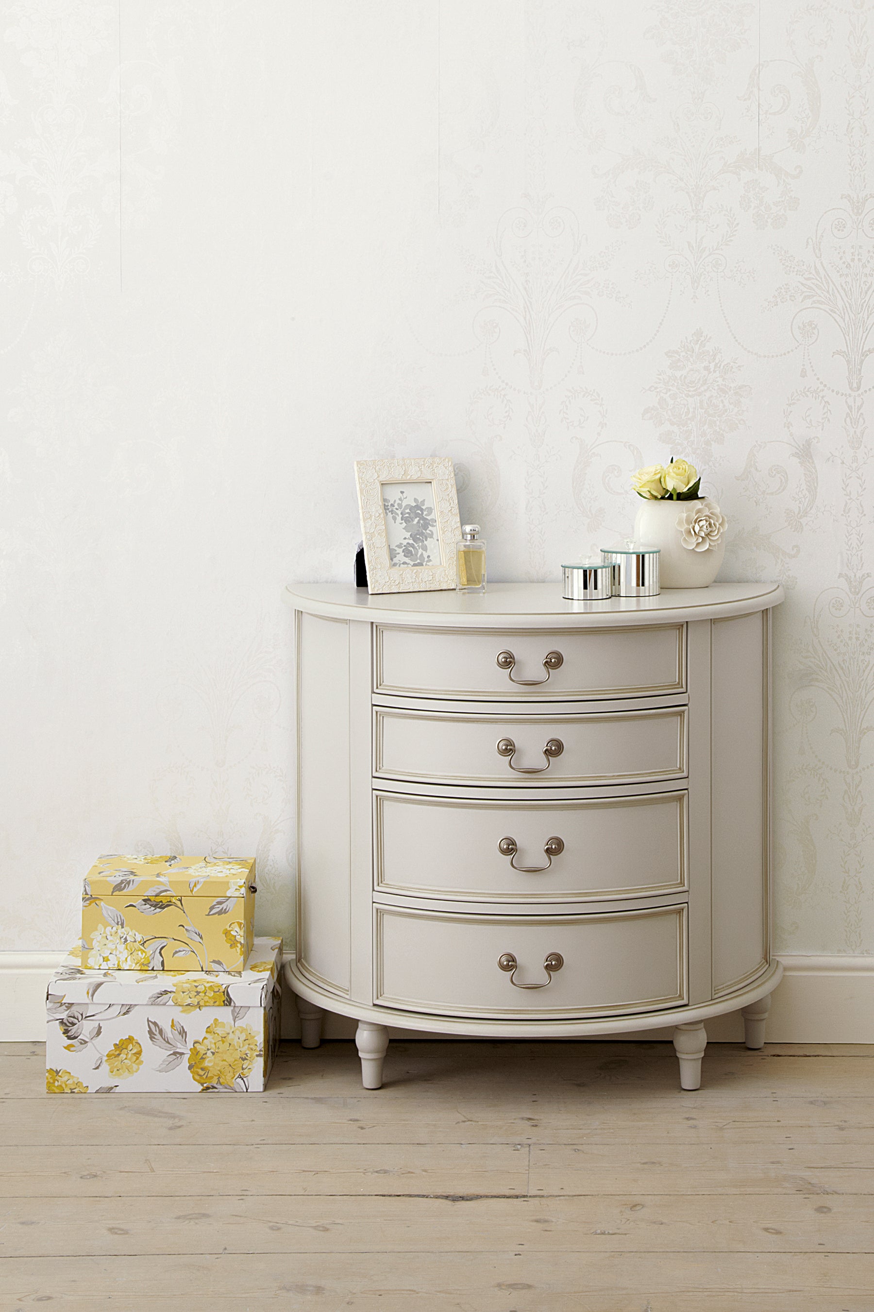 A dove grey 4-drawer half moon chest of drawers with a curved front and traditional metal handles with various ornaments sitting on top and next to it
