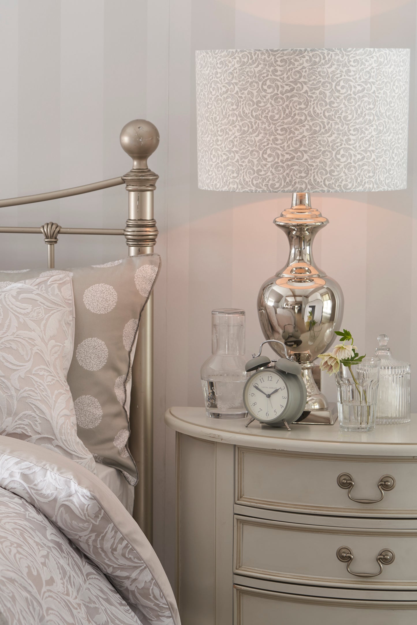 A dove grey 4-drawer half moon chest of drawers with a curved front and traditional metal handles next to a bed. A table lamp and ornaments sit on top
