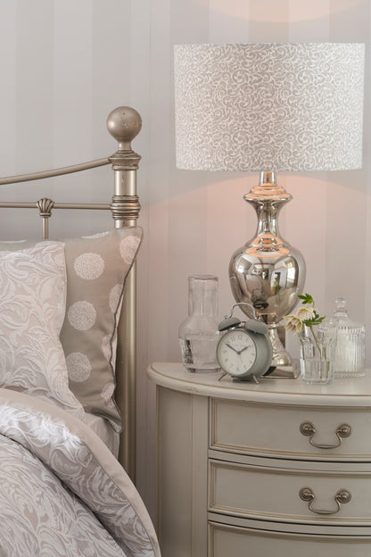 A dove grey 4-drawer half moon chest of drawers with a curved front and traditional metal handles next to a bed. A table lamp and ornaments sit on top