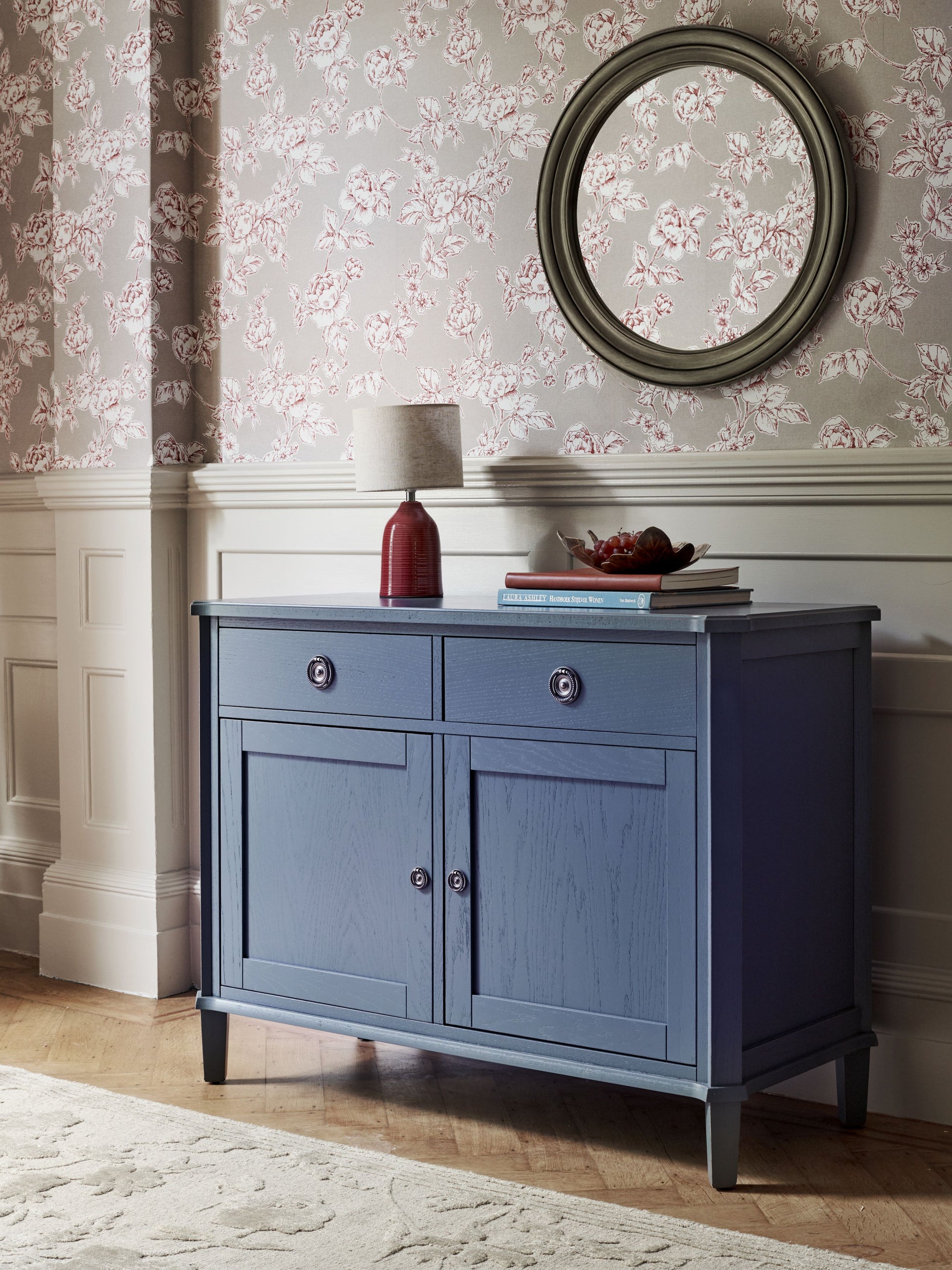 Blue sideboard against a floral wallpapered wall with a round mirror above.