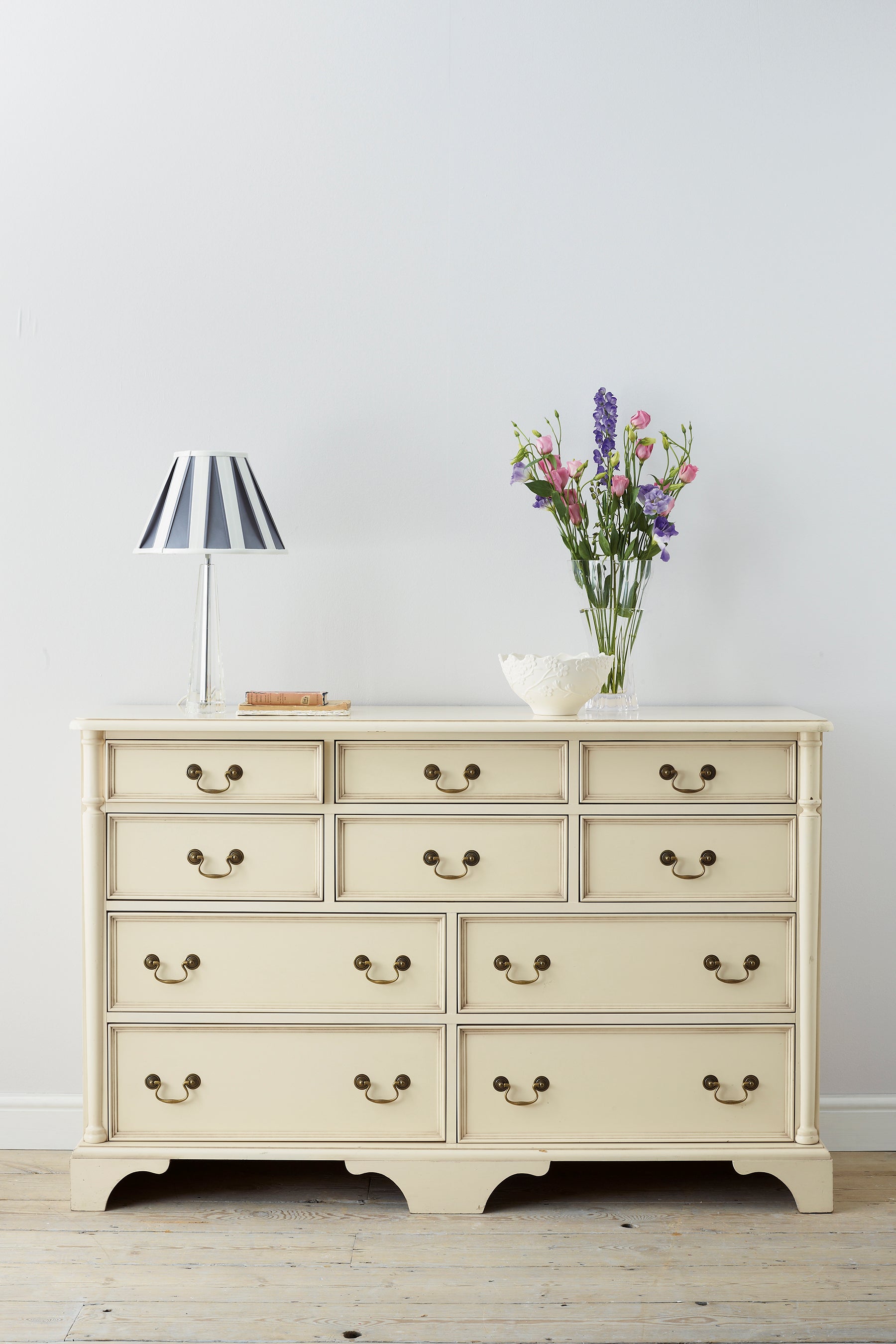 Beige dresser with drawers, lamp, and vase of flowers on a wooden floor.
