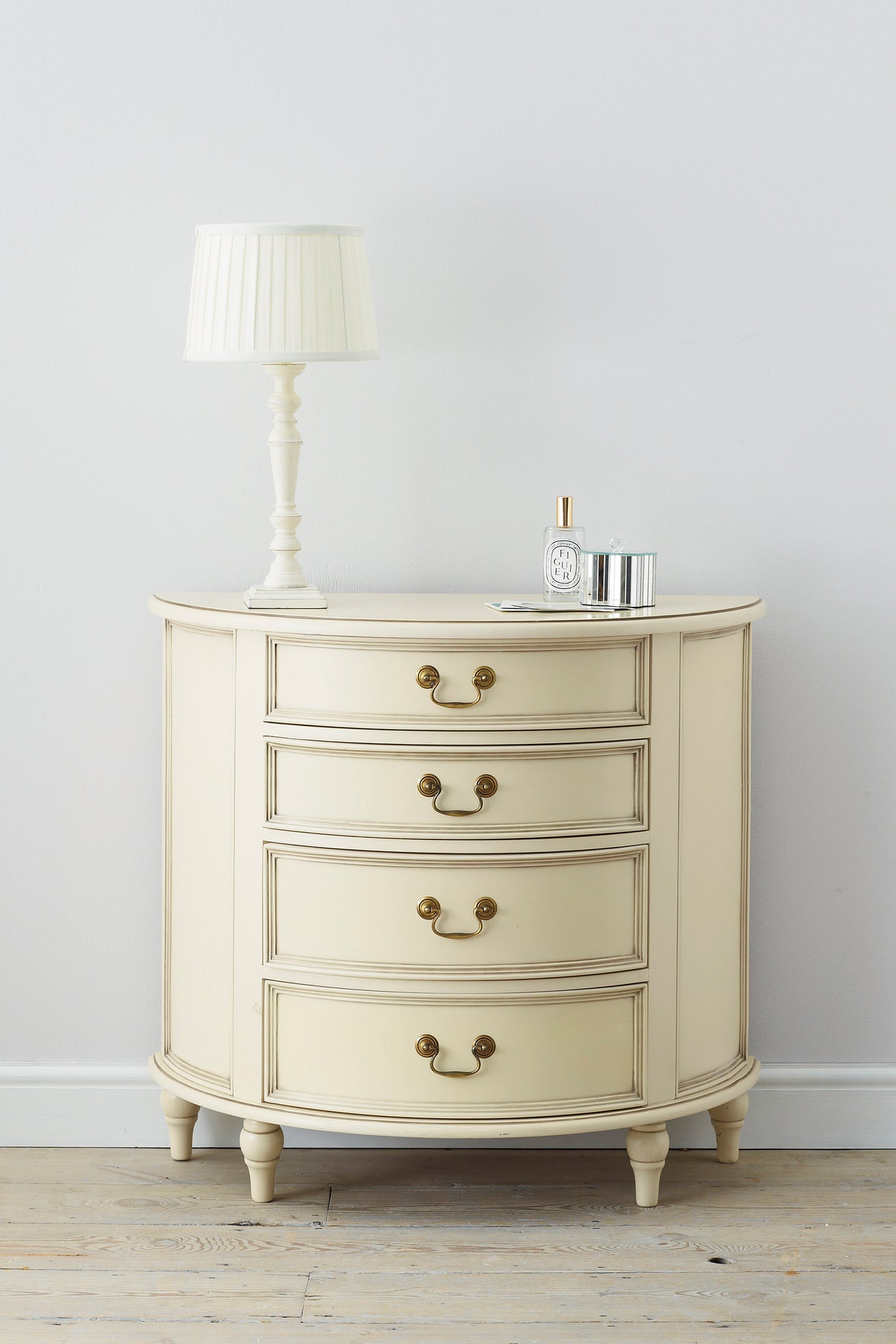 A front-facing view of an Ivory coloured half moon chest with four drawers and vintage styling. A lamp and some ornamental containers are shown to stand on top