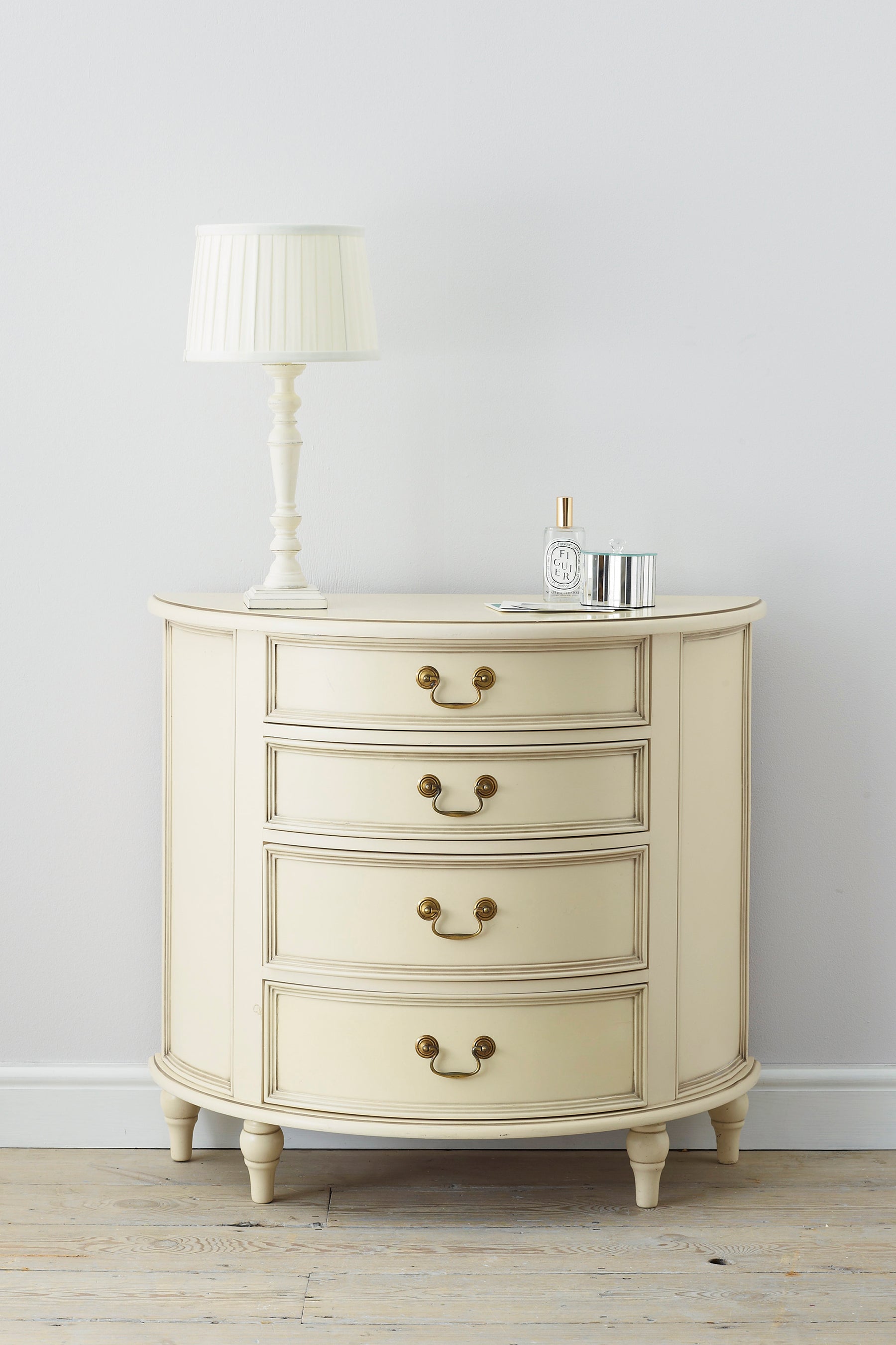 A front-facing view of an Ivory coloured half moon chest with four drawers and vintage styling. A lamp and some ornamental containers are shown to stand on top