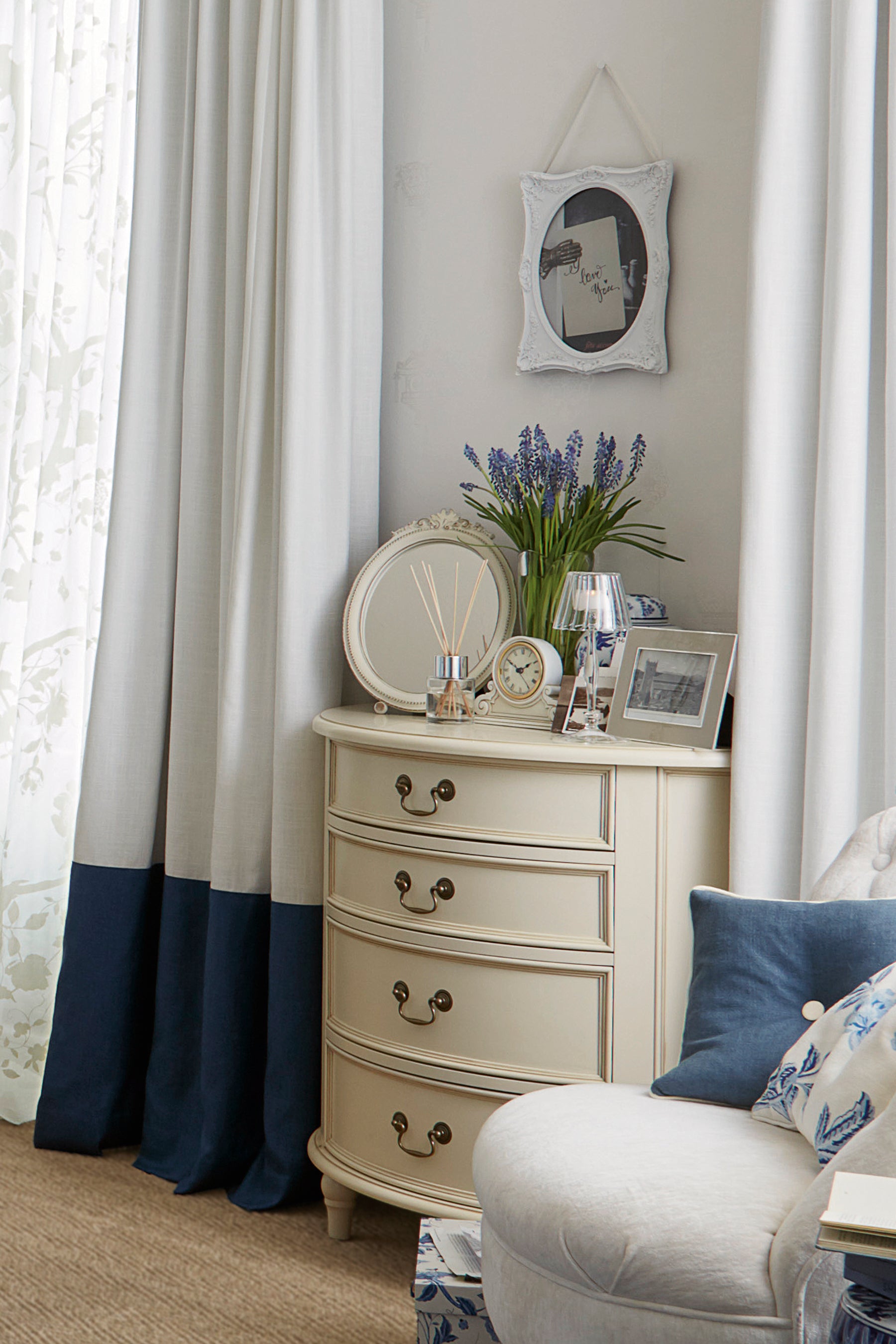 An Ivory coloured half moon chest with four drawers and vintage styling with plenty of ornaments including a reed diffuser, clock, mirror, picture frame and vase of flowers