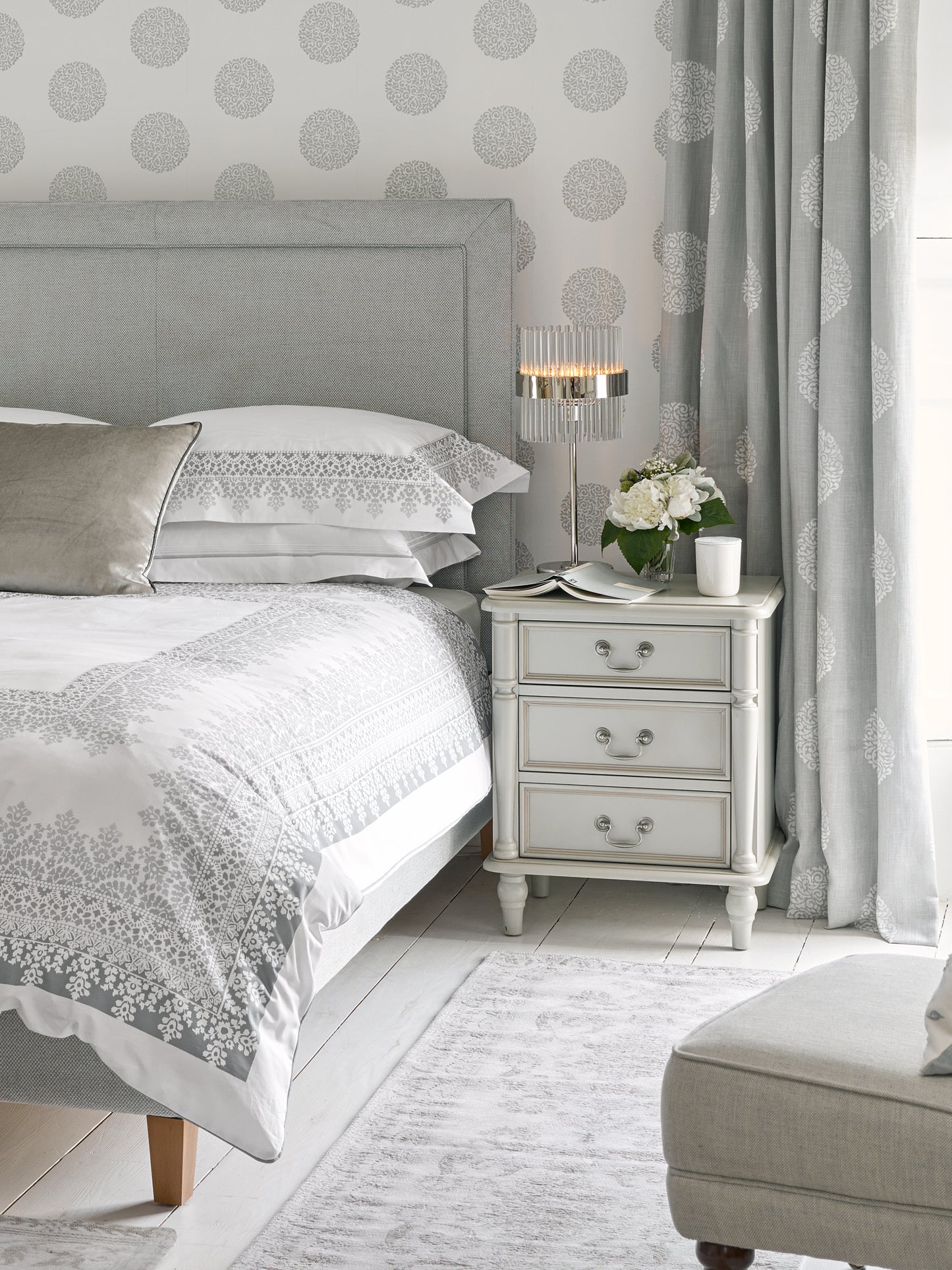 A dove grey 3-drawer bedside chest with traditional design and metal handles next to a bed with additional accessories
