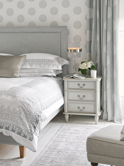 A dove grey 3-drawer bedside chest with traditional design and metal handles next to a bed with additional accessories