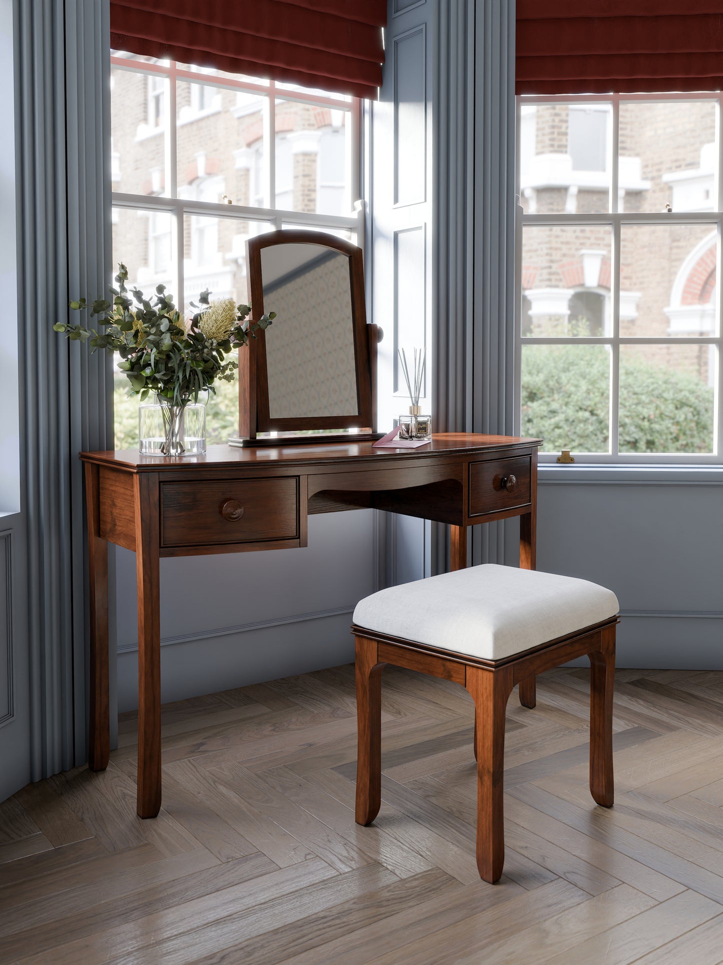 A A traditional dark wooden 2-drawer dressing table with a mirror and a stool. The stool has a white seat pad. The set is standing against a blue wall with large windows
