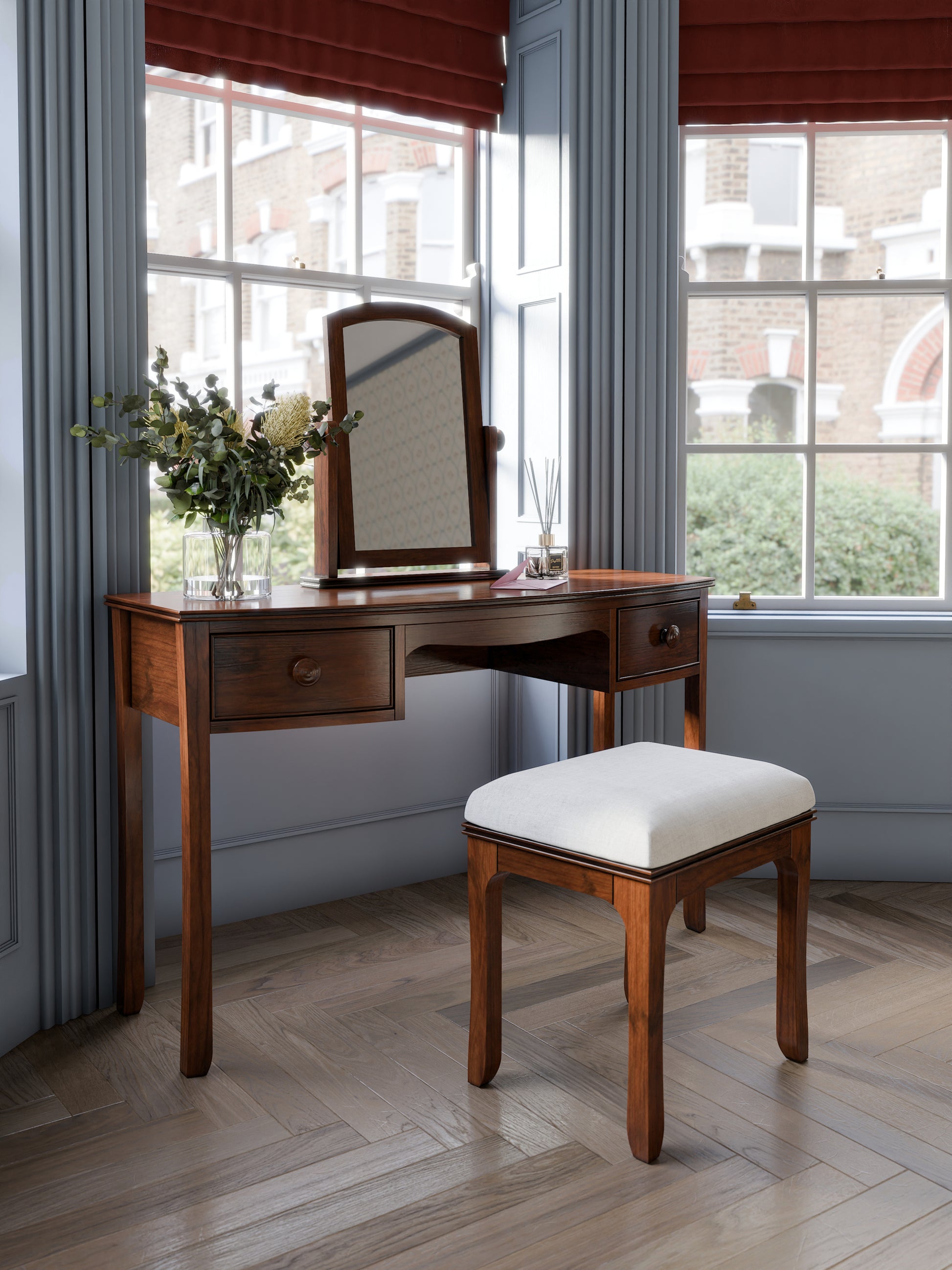 A A traditional dark wooden 2-drawer dressing table with a mirror and a stool. The stool has a white seat pad. The set is standing against a blue wall with large windows