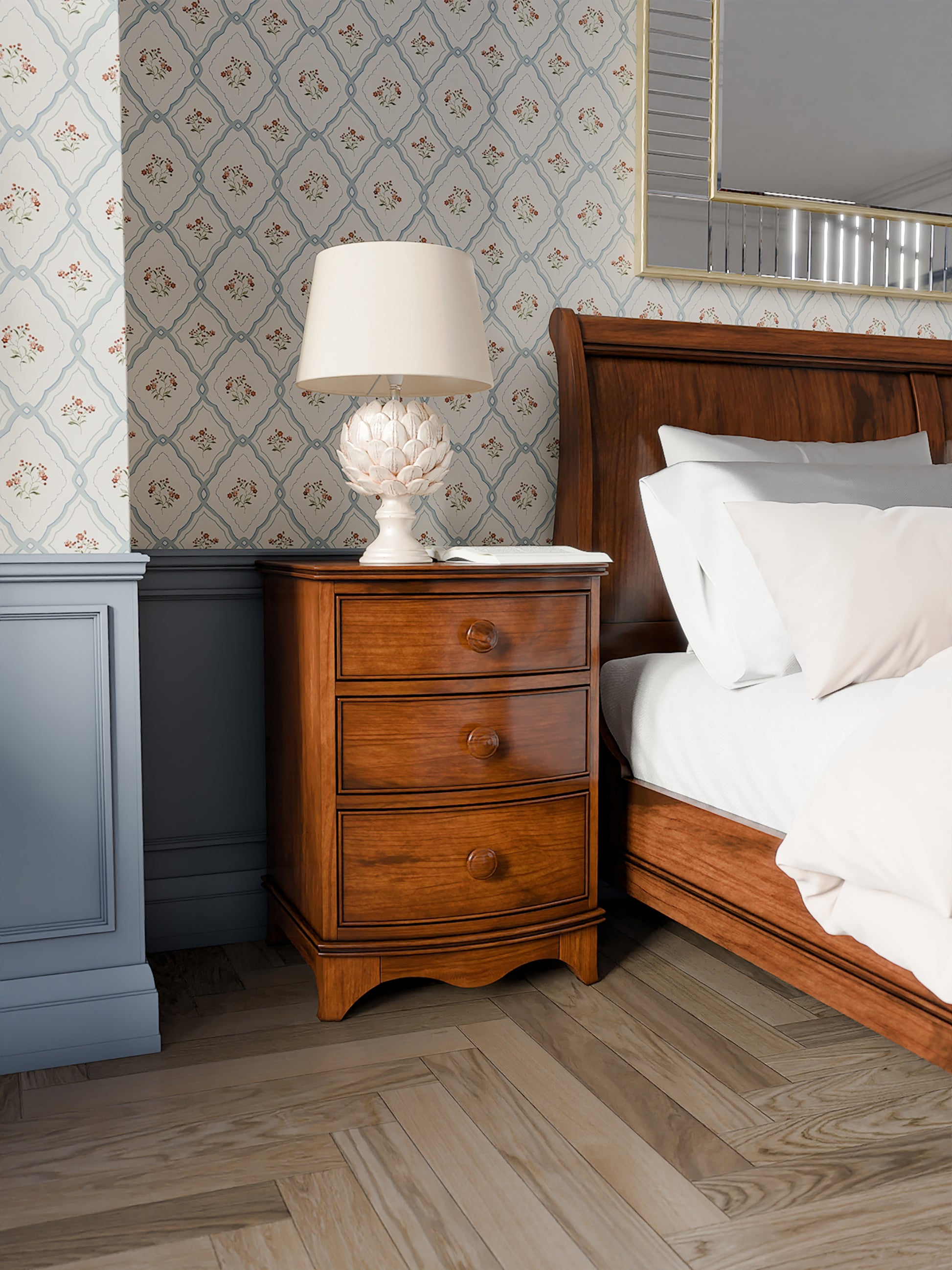 A dark wooden 3-drawer bedside chest with a curved front and traditional design, a white table lamp on top and standing next to a dark wooden bed with white bed linen