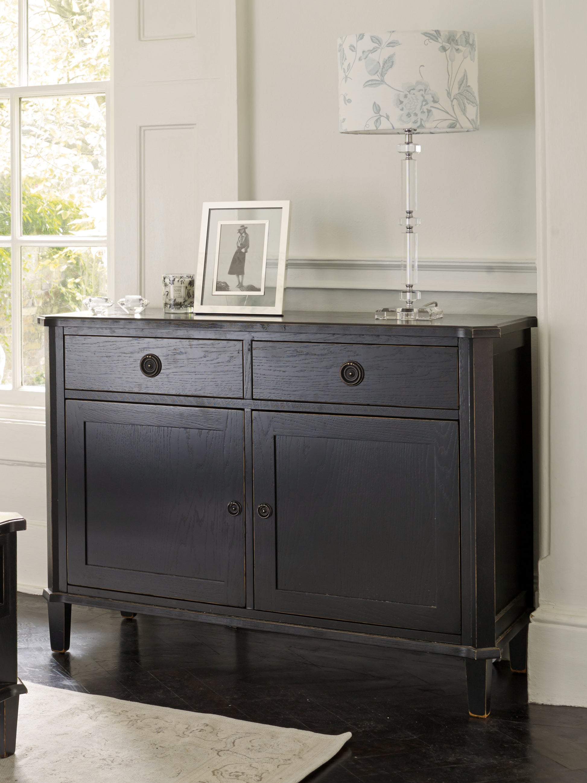Dark wooden sideboard with a lamp and picture frame in a room with large windows.
