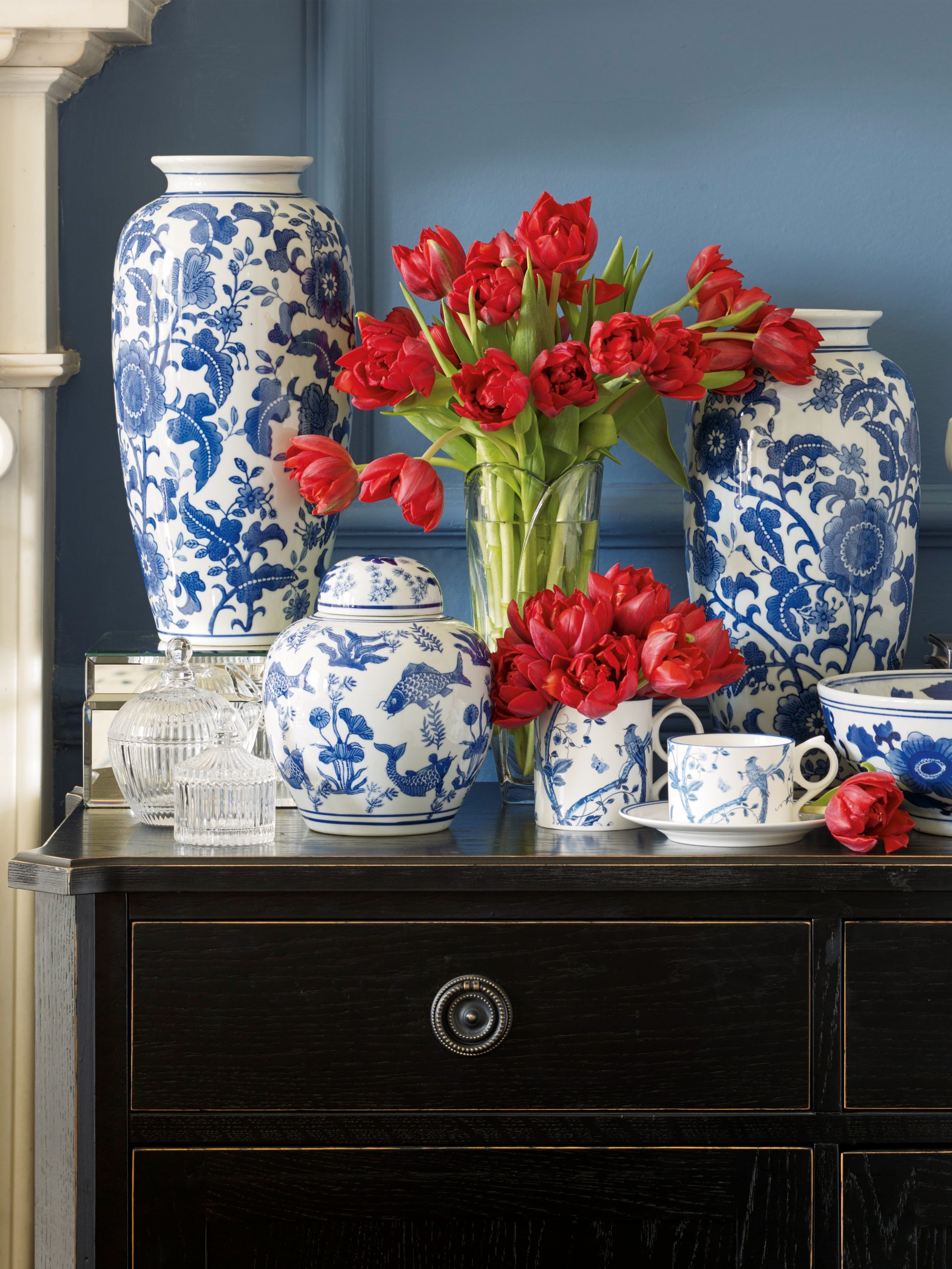 Decorative setting with blue and white vases, a teapot, and red tulips on a dark surface against a blue wall.