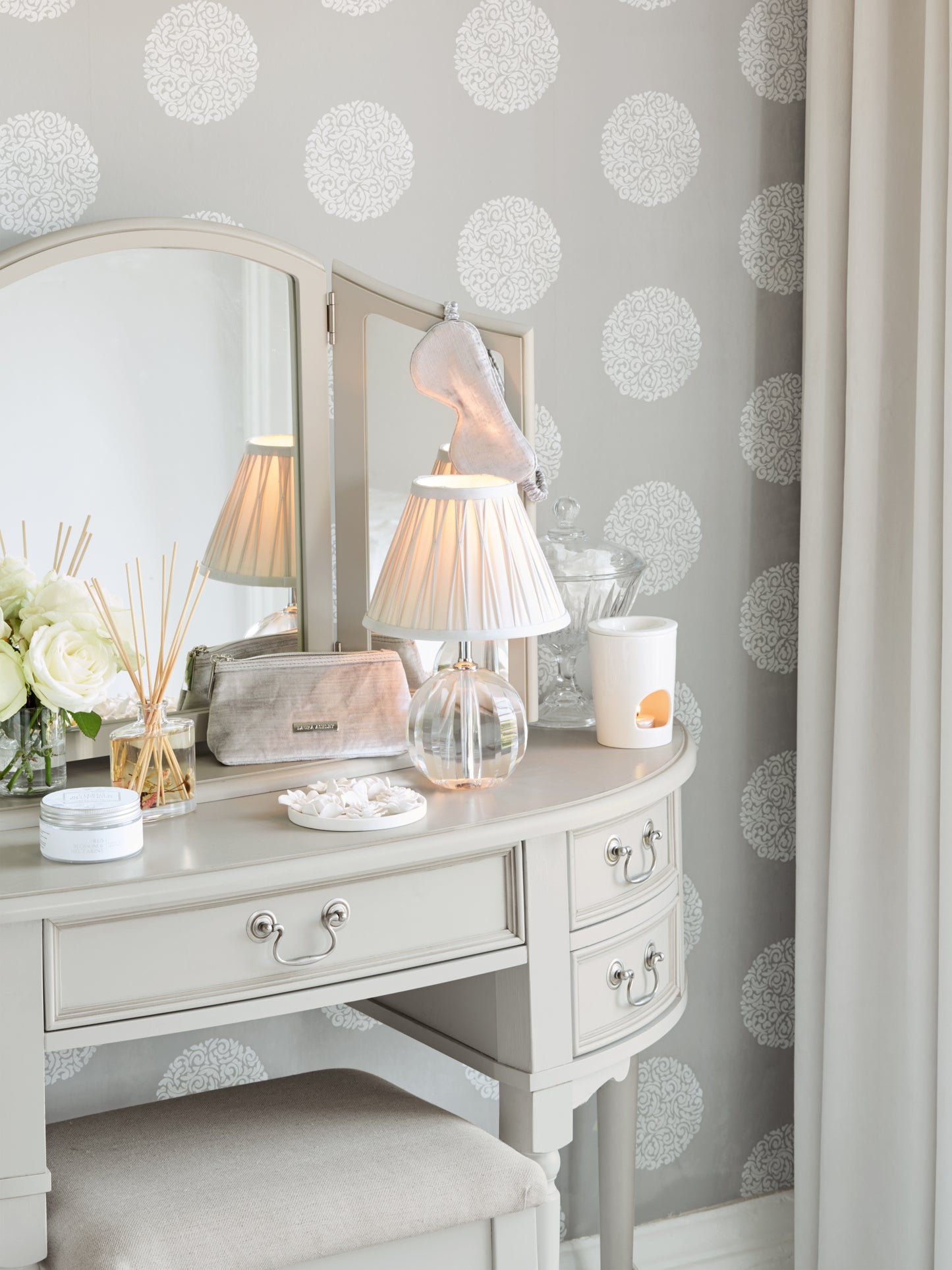 A Clifton Dove Grey 5 Drawer Dressing Table with a stool set in front of it. The table is large with multiple drawers and the stool is cushioned with various accessories including a table lamp, some flowers and a reed diffuser