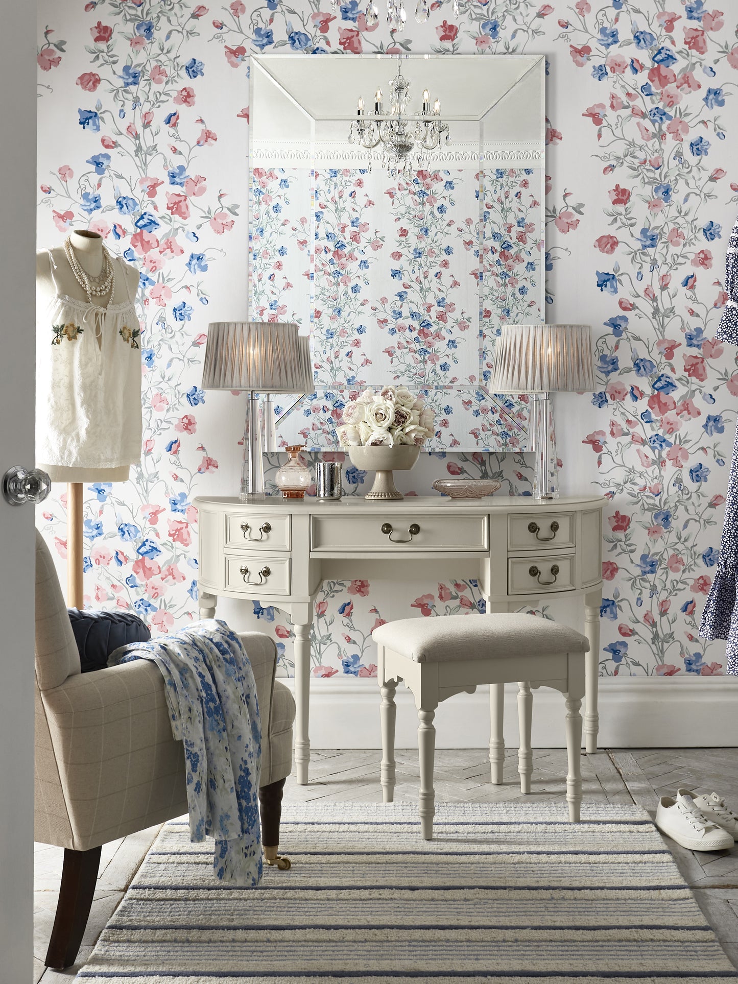 A Clifton Dove Grey 5 Drawer Dressing Table with a stool set in front of it. The table is large with multiple drawers and the stool is cushioned with various ornaments and accessories including an armchair next to the table, 2 table lamps and a floral wallpaper background