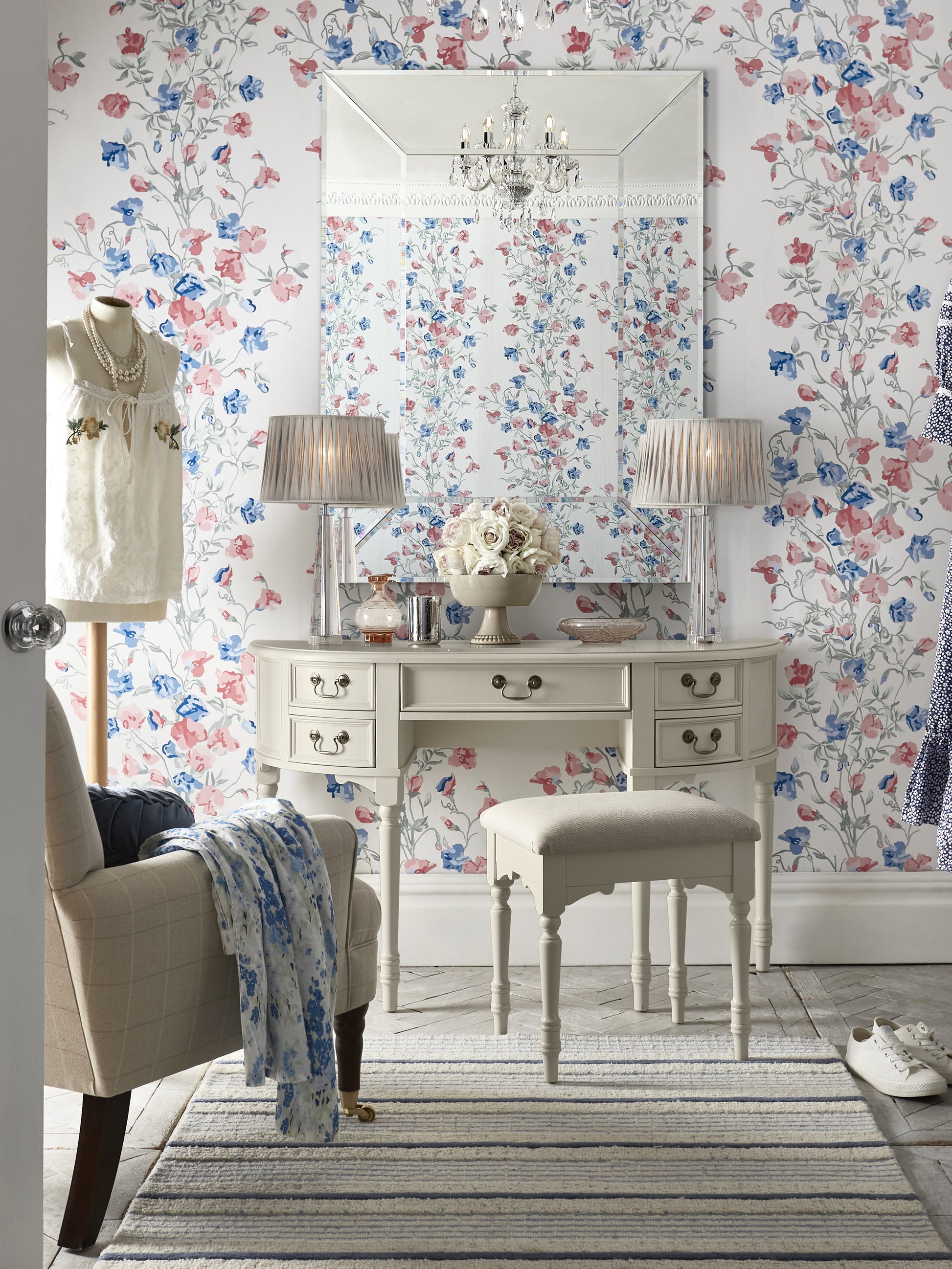 A Clifton Dove Grey 5 Drawer Dressing Table with a stool set in front of it. The table is large with multiple drawers and the stool is cushioned with various ornaments and accessories including an armchair next to the table, 2 table lamps and a floral wallpaper background