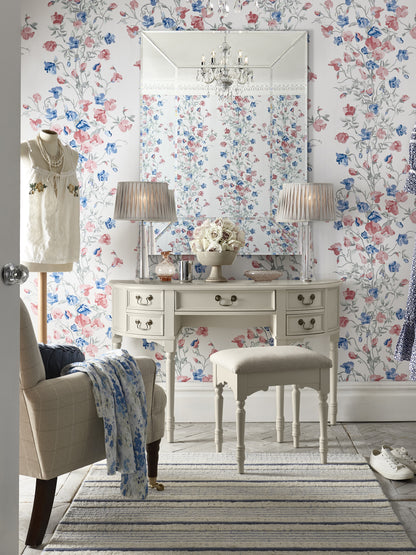 A Clifton Dove Grey 5 Drawer Dressing Table with a stool set in front of it. The table is large with multiple drawers and the stool is cushioned with various ornaments and accessories including an armchair next to the table, 2 table lamps and a floral wallpaper background