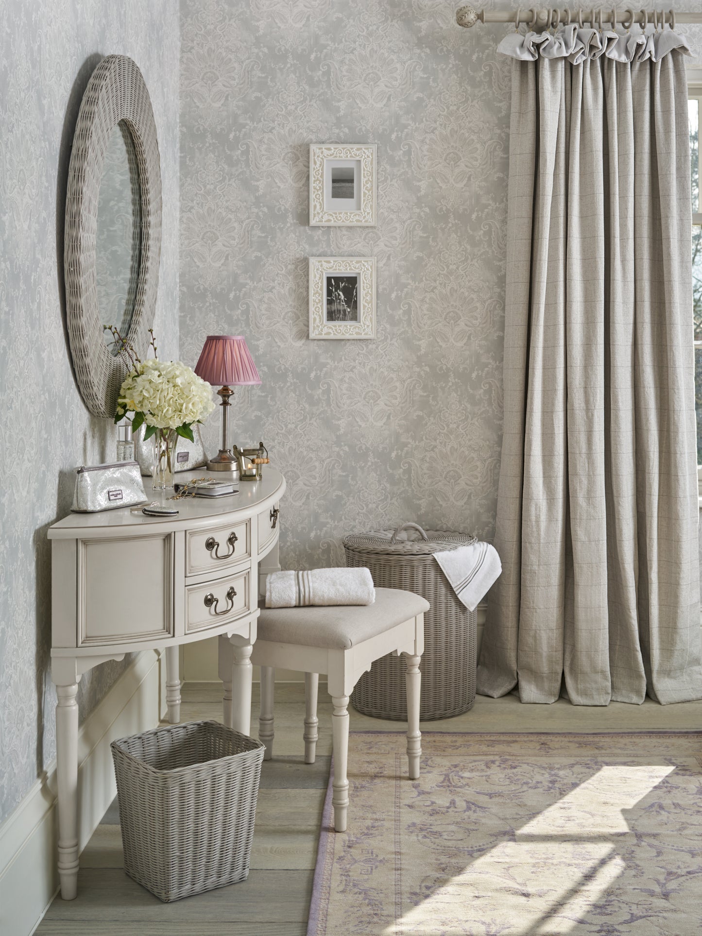 A Clifton Dove Grey 5 Drawer Dressing Table with a stool set in front of it. The table is large with multiple drawers and the stool is cushioned and show various ornaments and accessories.
