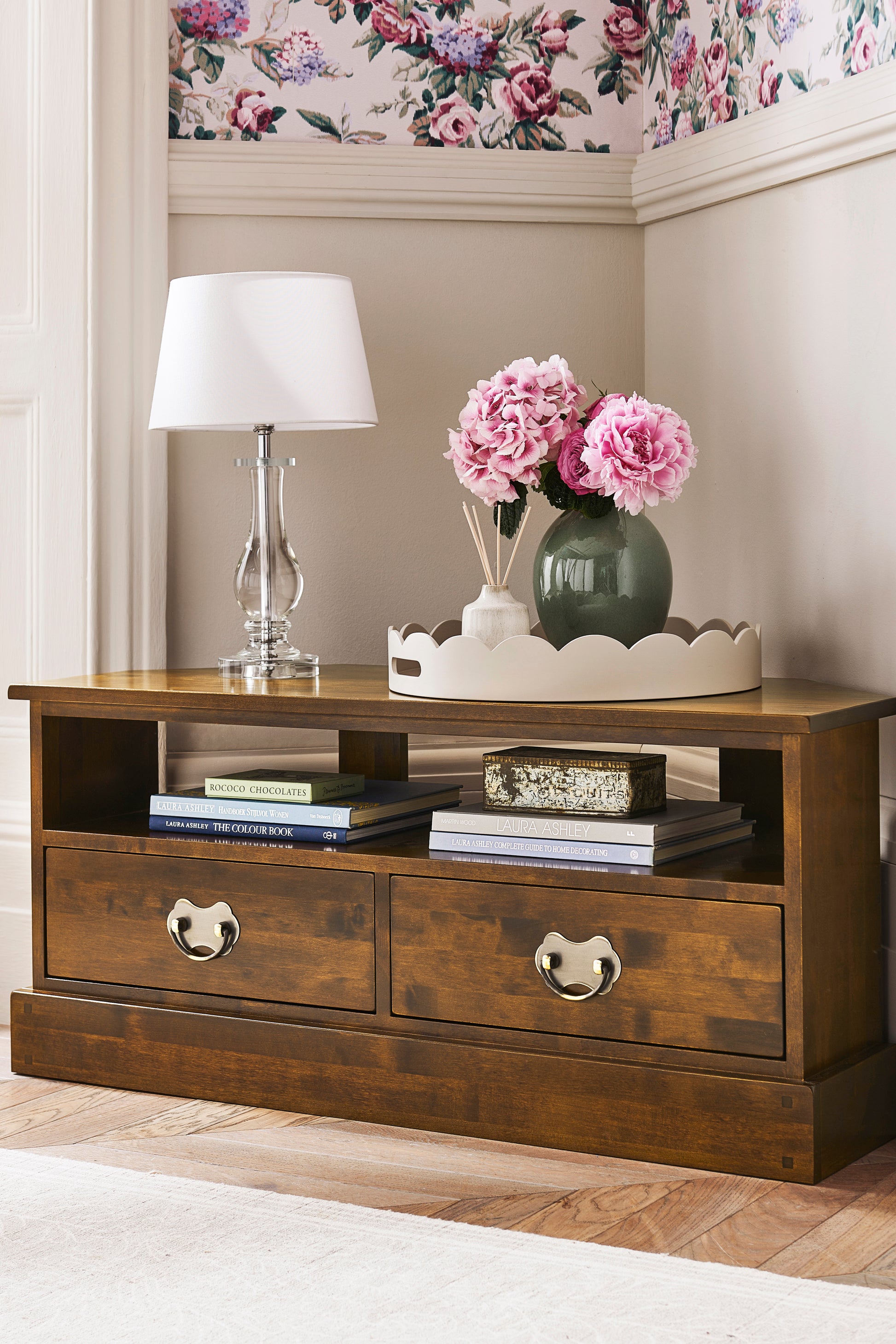 Wooden side table with drawers, lamp, and flowers in a stylish room.