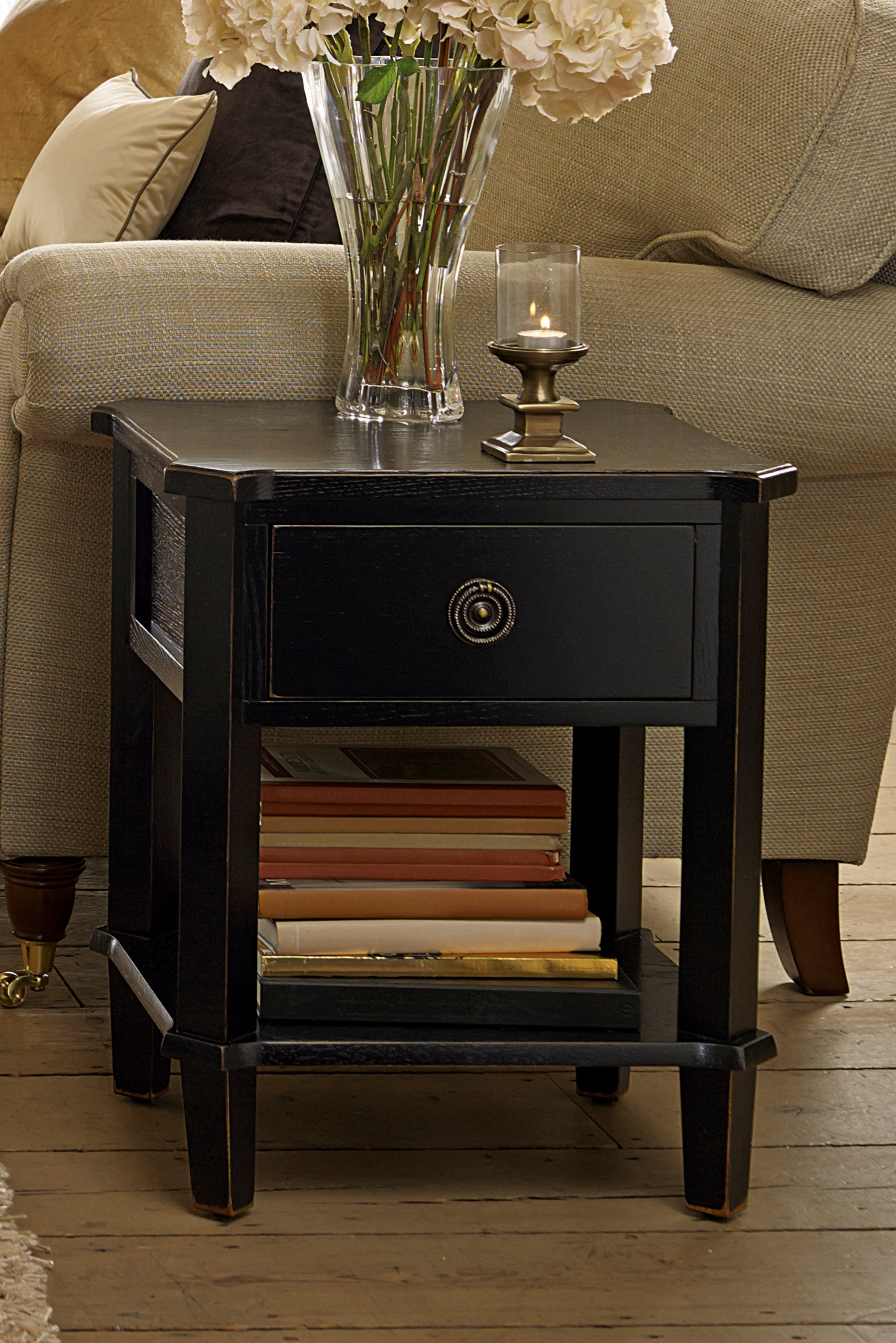 Black end table with a drawer, candle, and vase on a wooden floor next to a beige sofa.