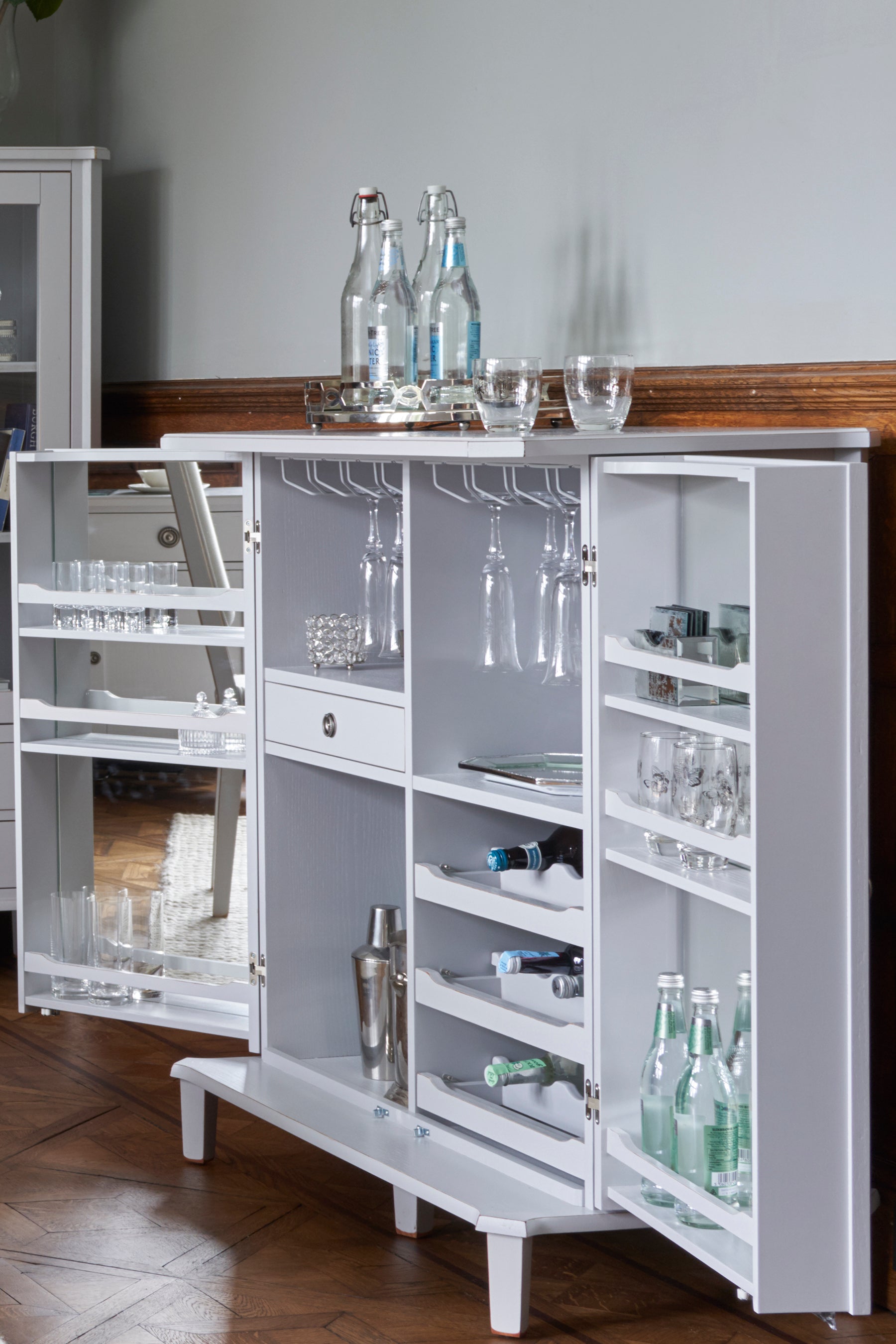 Pale grey drinks cabinet with glass shelves and storage compartments in a room setting.