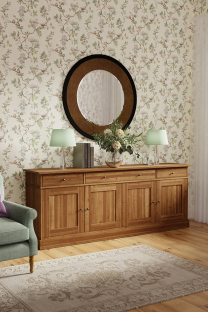 Honey coloured wooden sideboard with mirror and lamps in a room with floral wallpaper.