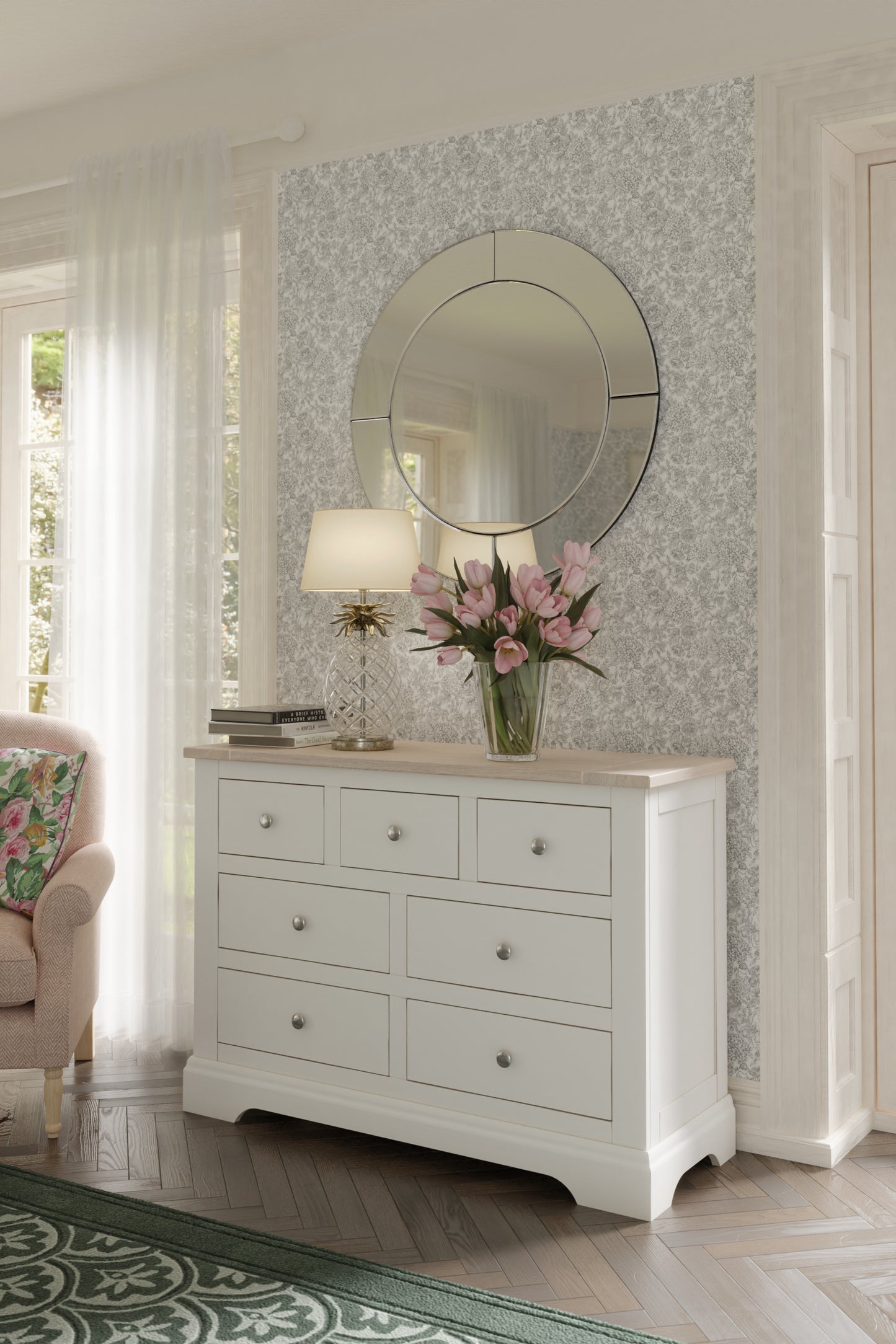 A white Laura Ashley Dorset 3+4 Drawer Chest with a wooden top against a floral background. A table lamp, some books and flowers lay on top.