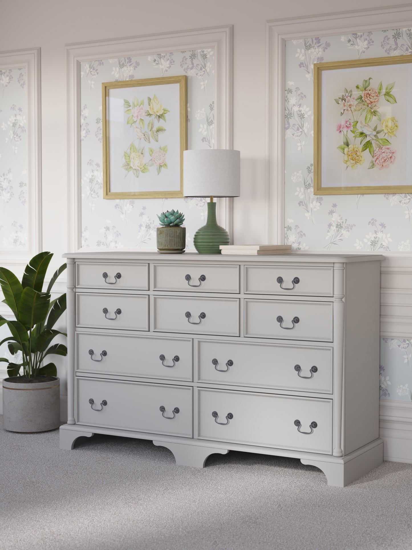 A dove grey Laura Ashley Clifton 6+4 drawer chest with traditional design against a floral background with a house plant next to it, a table lamp on top with various ornaments