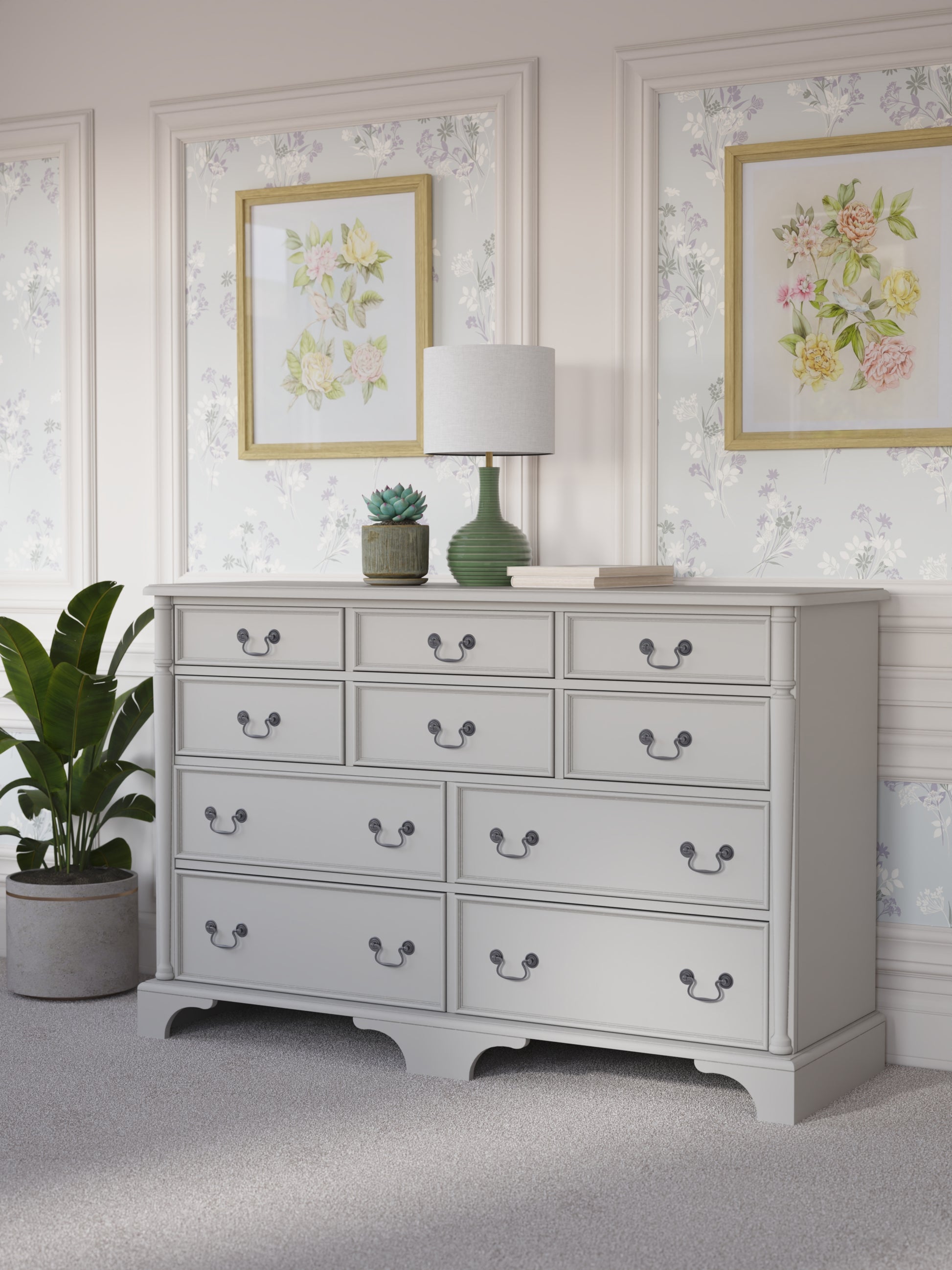A dove grey Laura Ashley Clifton 6+4 drawer chest with traditional design against a floral background with a house plant next to it, a table lamp on top with various ornaments